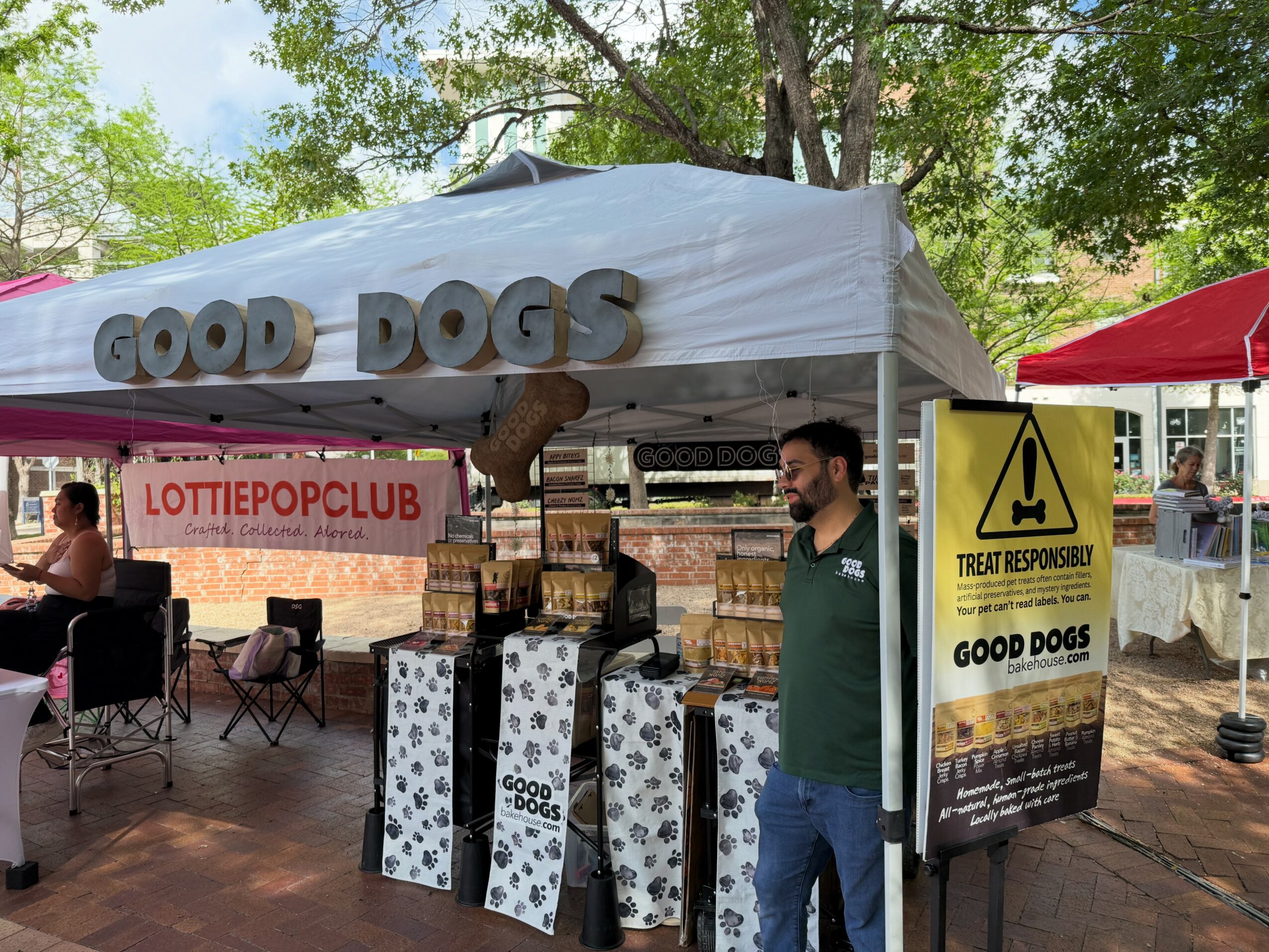 Outdoor event booth with a white tent labeled 'Good Dogs,' people sitting nearby, and a yellow sign about treat responsibility.