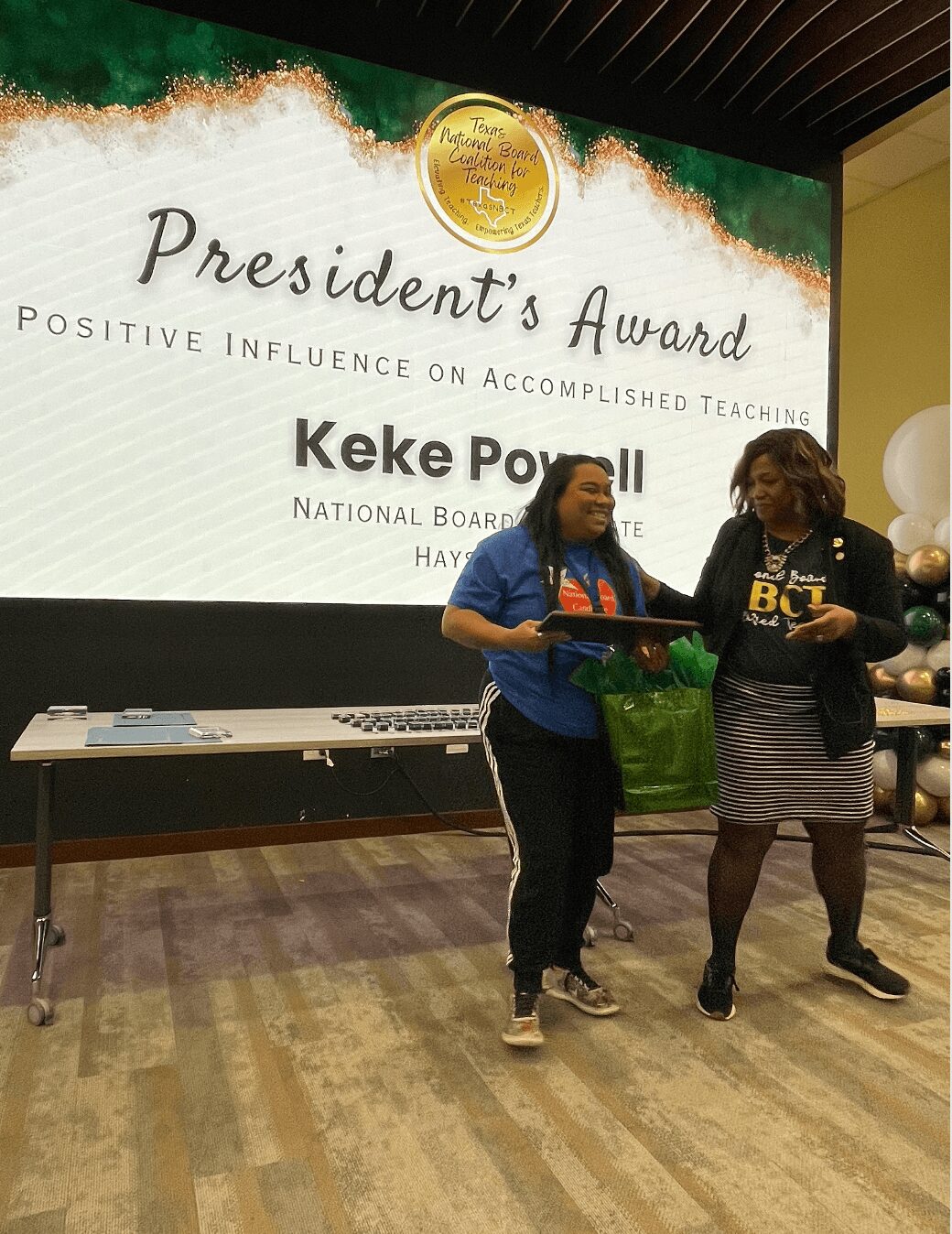Two women standing in front of a large screen with a President's Award presentation, one holding a green bag, smiling.