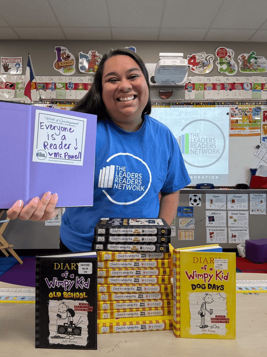 Smiling person in blue shirt behind table with books and a sign in a classroom.