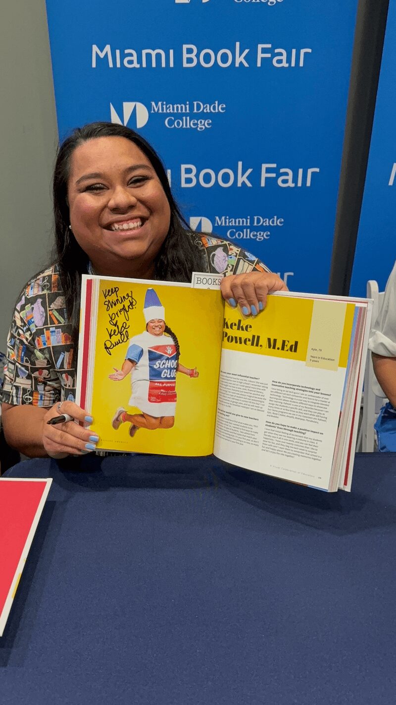 Smiling woman holding open book at book fair, background shows blue banner with white text, table in front.