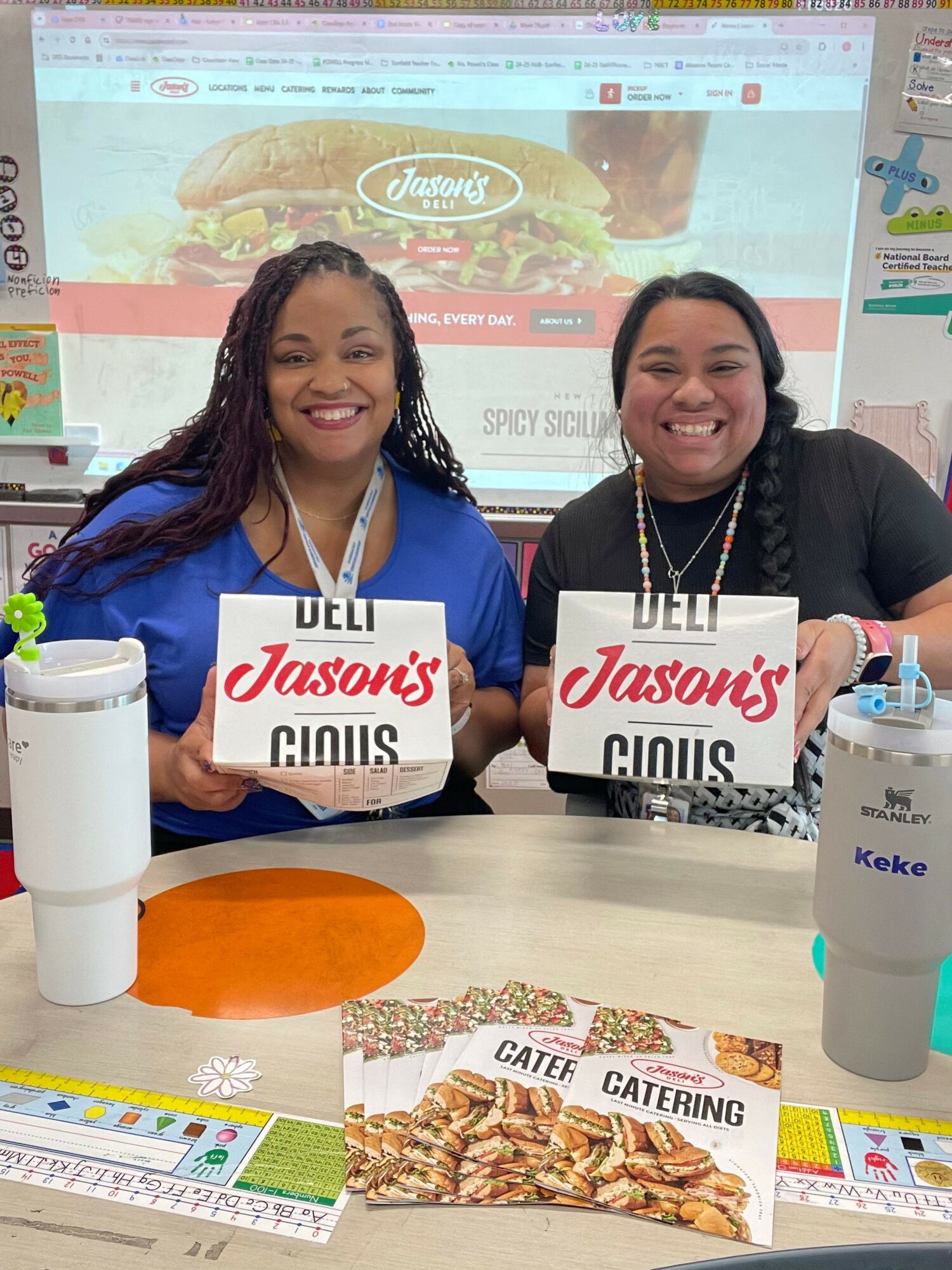 Two women sitting at a table holding Jayson’s Hills signs, with a promotional display and food items on the table.