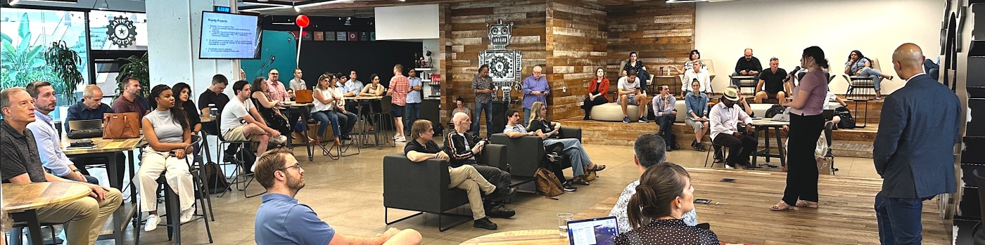 People seated at tables and chairs in a large room with a presentation screen and wooden wall, some standing and talking.