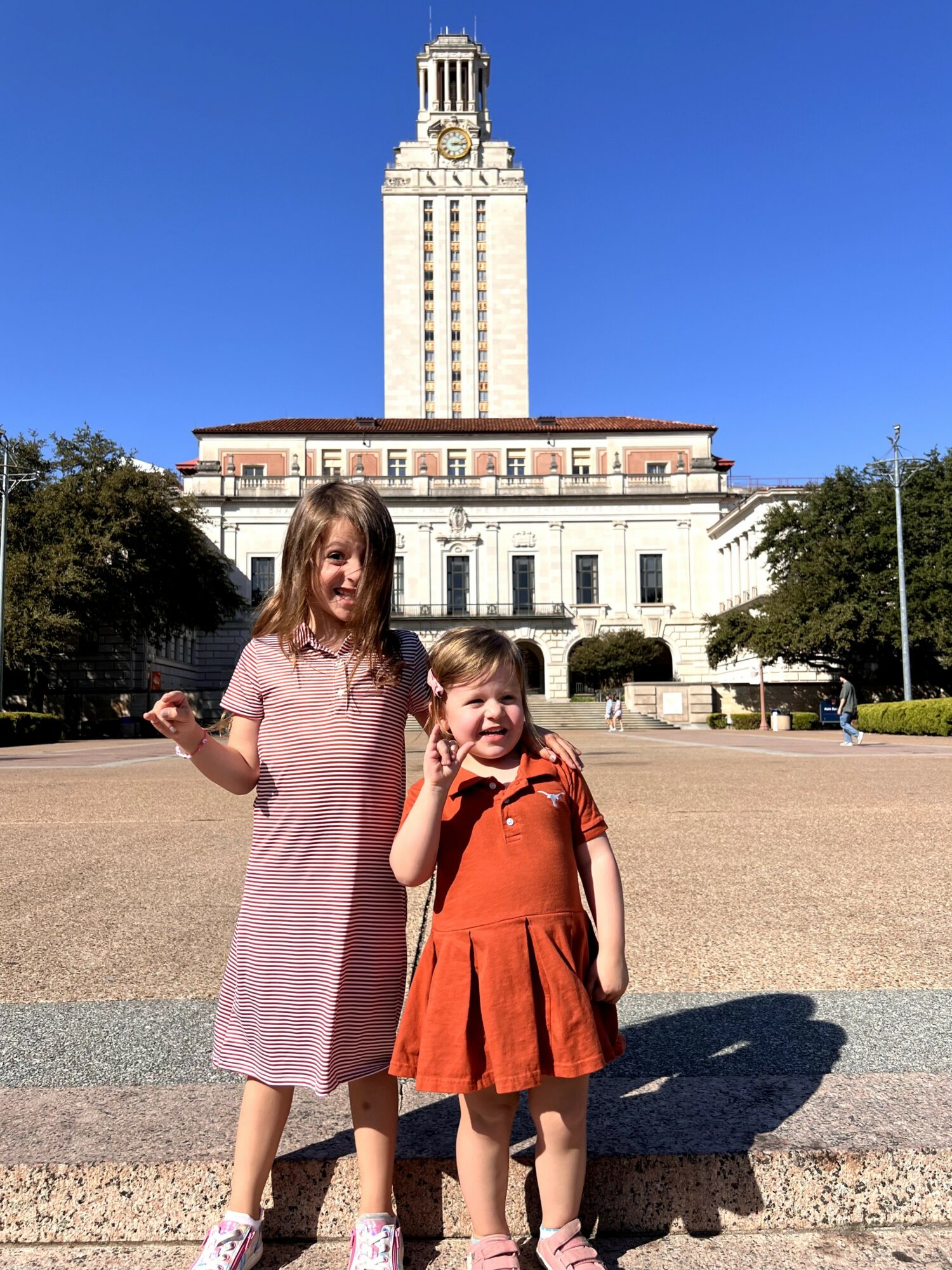 Two children standing outdoors in front of a tall building with a clock tower, clear blue sky, and trees in the background.