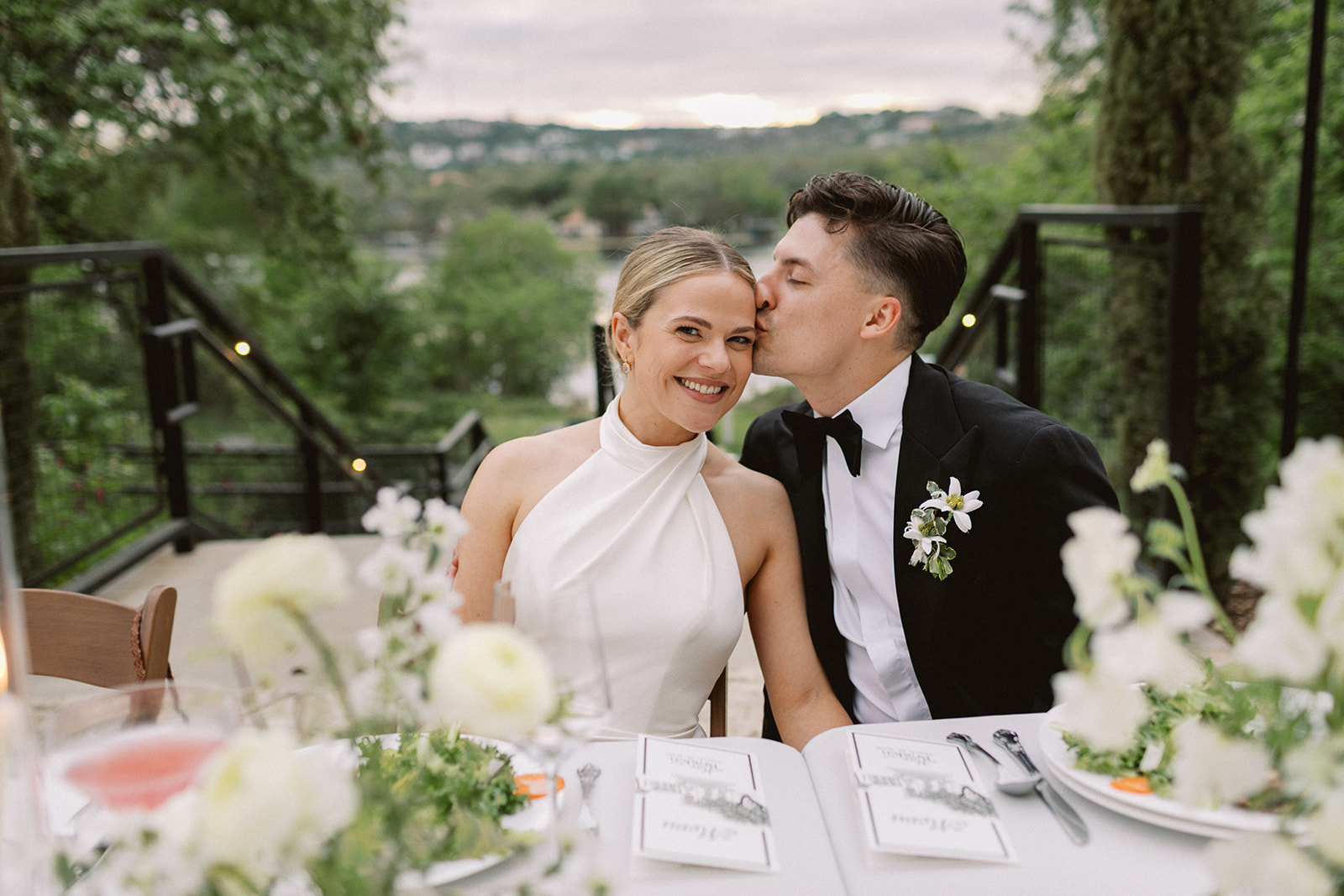 A bride and groom sit at a table outdoors, smiling and sharing a kiss, with floral decorations and greenery in the background.