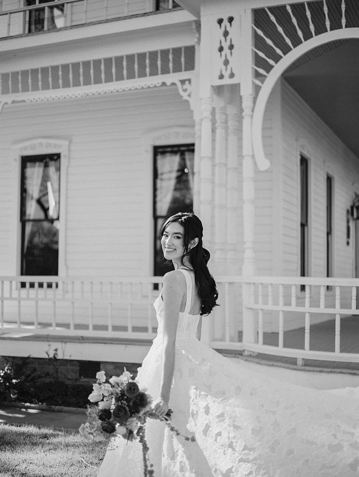 Woman in wedding dress holding bouquet, standing outside a house with decorative architecture, smiling, black and white photo.