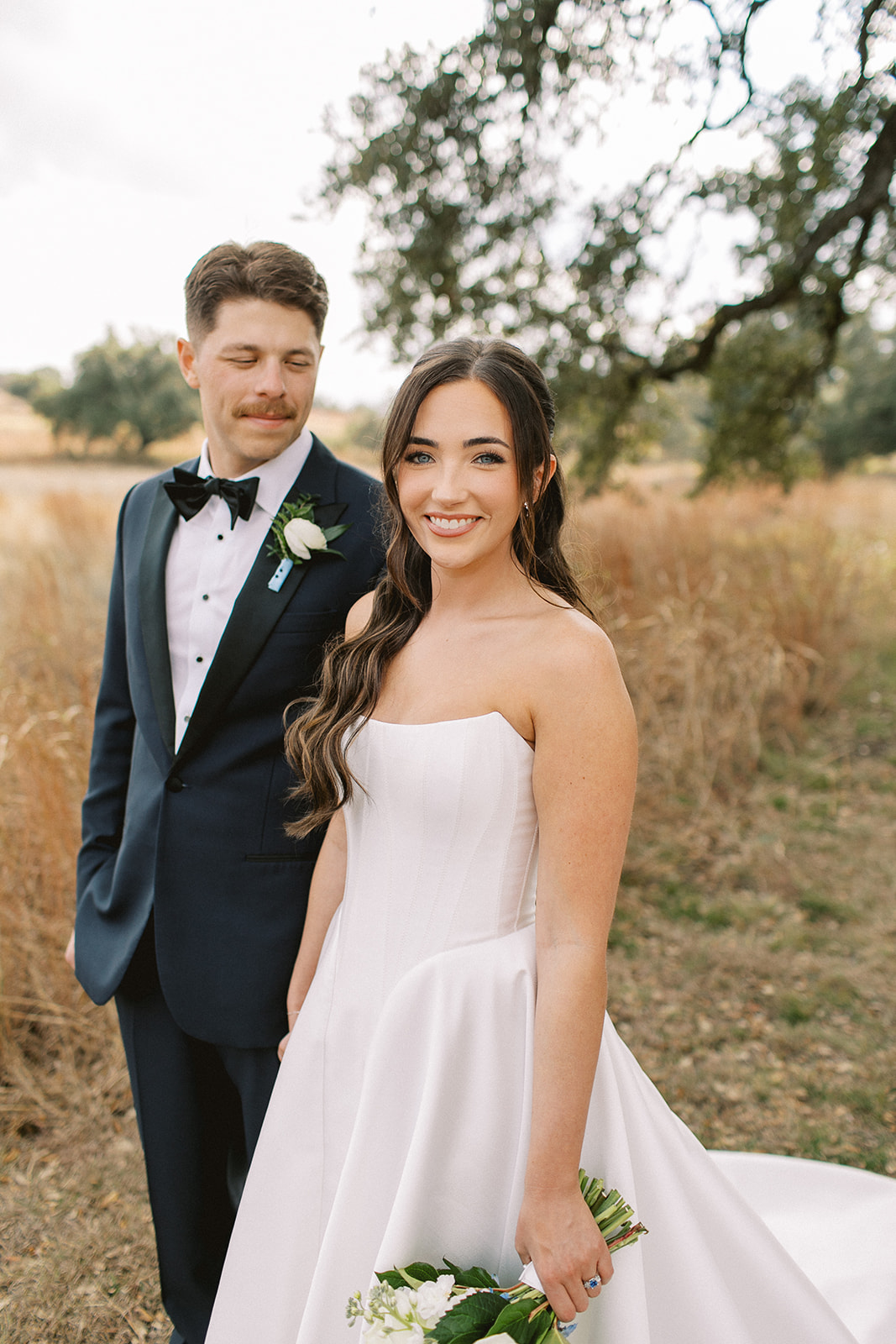 A smiling woman in a white strapless dress holding a small bouquet, standing outdoors with a man in a tuxedo behind her.