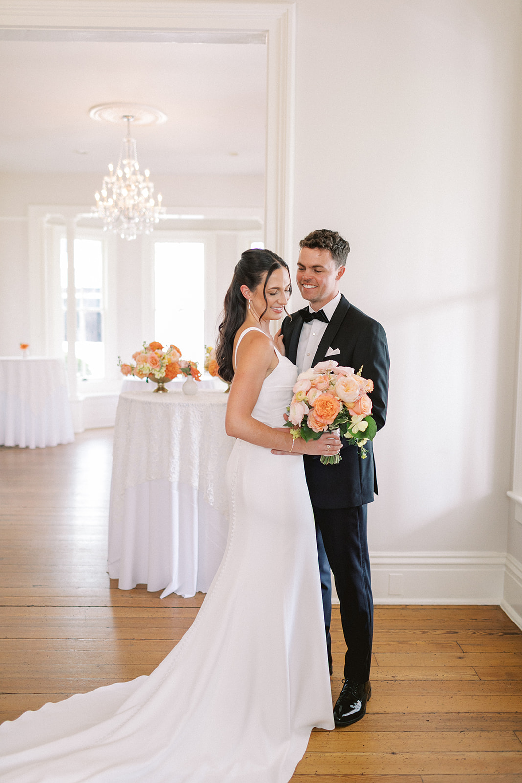 Bride and groom stand together indoors, bride holding a bouquet of pink and peach flowers, smiling at each other.