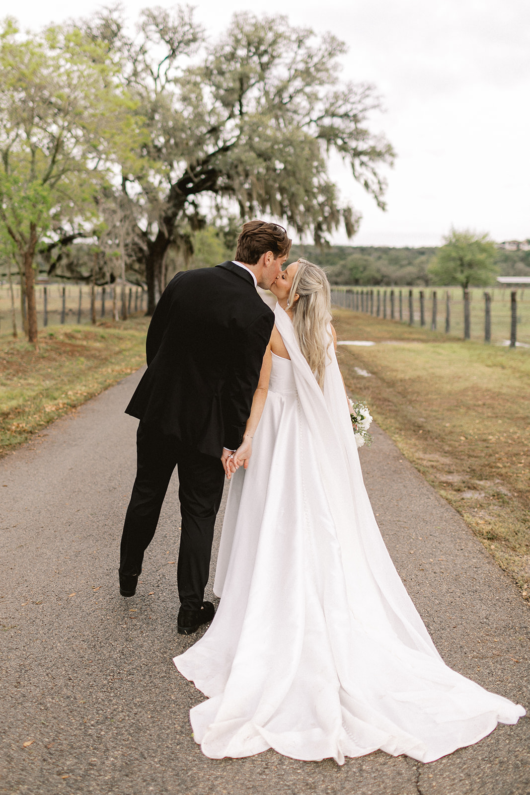 A bride and groom walk hand in hand on a rural path, with trees and a fence in the background, seen from behind.