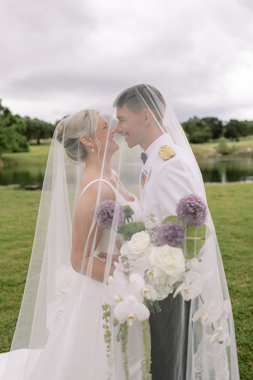 Bride and groom face each other under a veil outdoors, holding a bouquet of white and purple flowers, with a lake and trees in background.