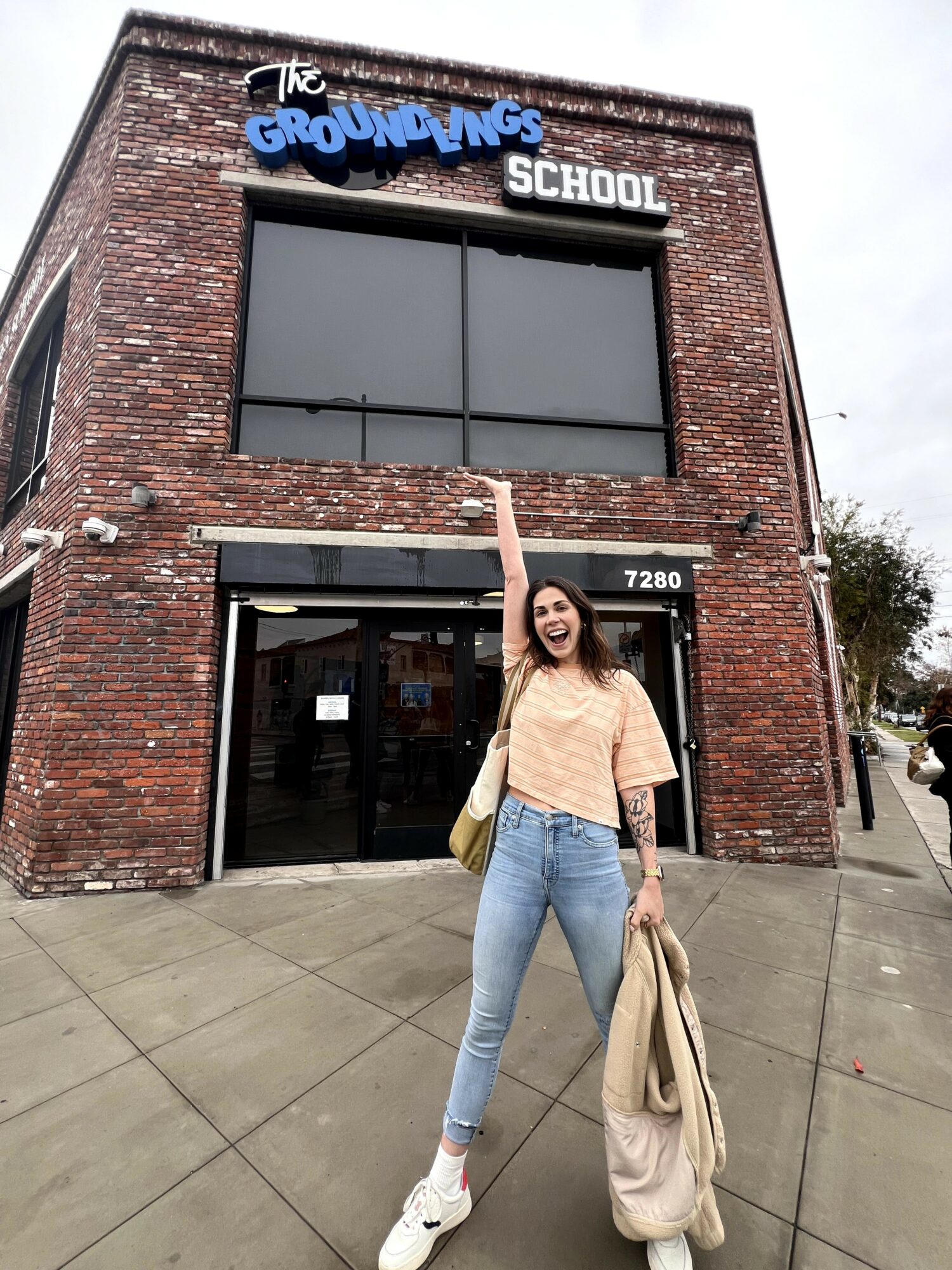 Young woman standing outside a brick school building, smiling, holding a coat, with a sign reading 'The Groundlings School'.