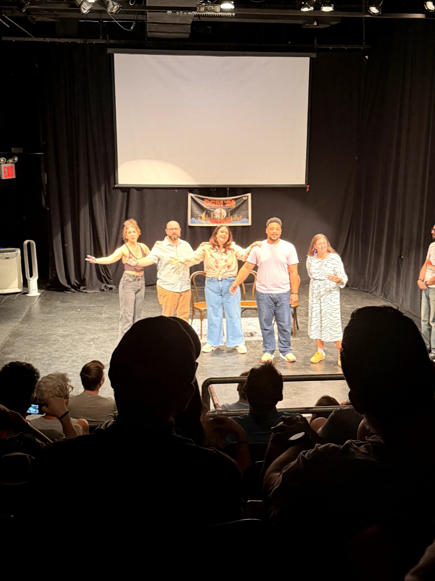 Six people holding hands on stage in front of an audience, with a large blank screen behind them.