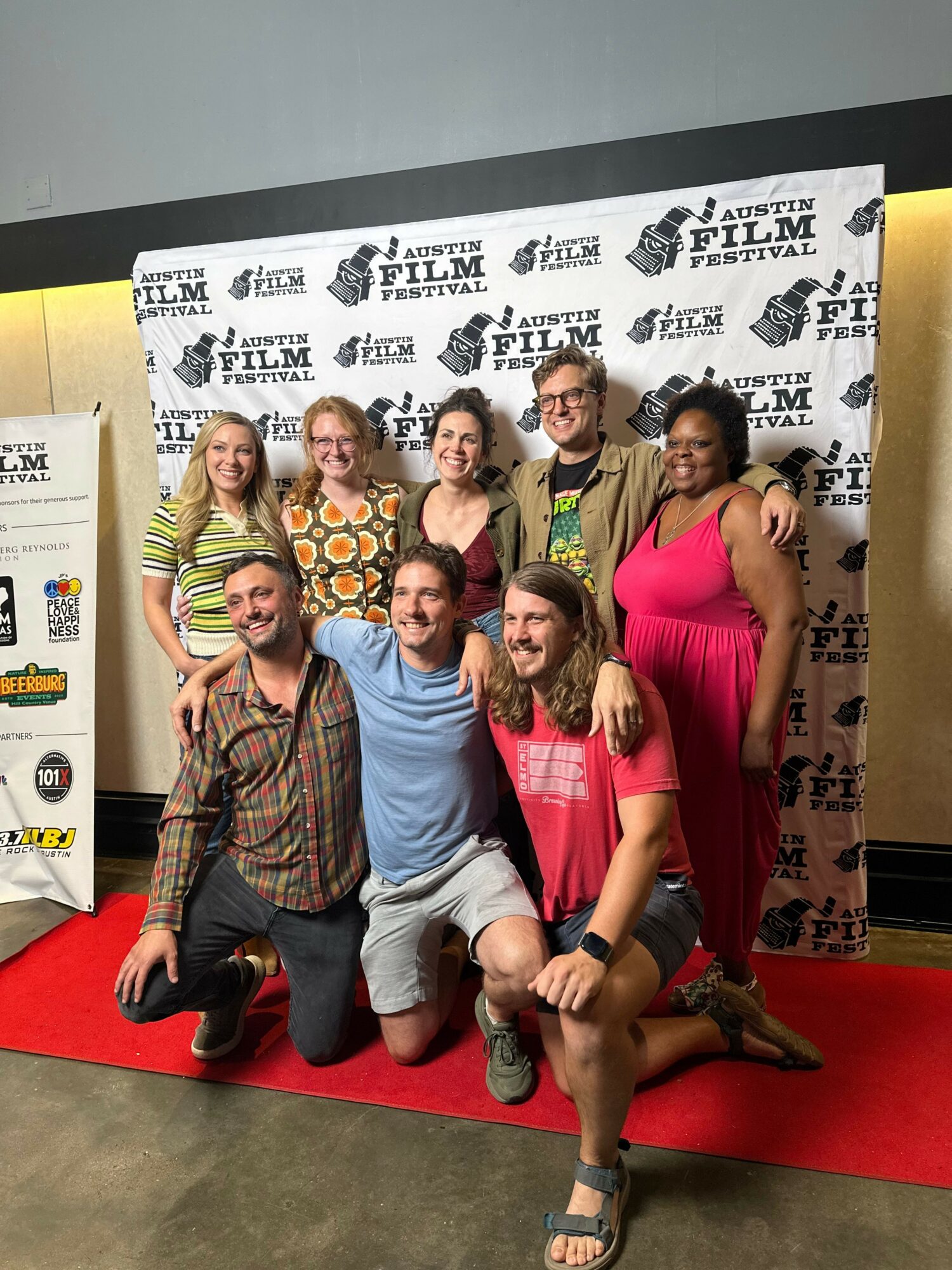 Group of people posing on a red carpet in front of a backdrop with logos, some sitting and some standing, smiling.