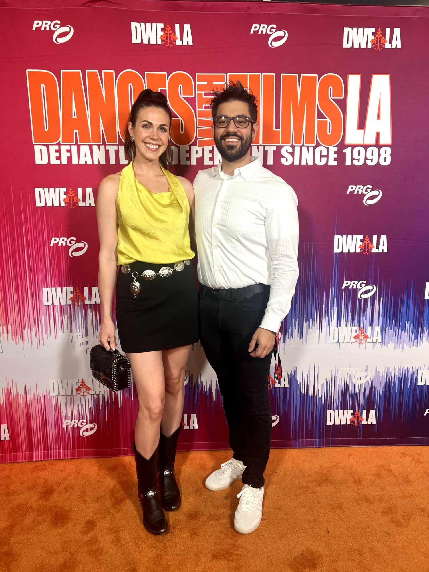 Two people standing on an orange carpet in front of a red backdrop with event text, smiling at the camera.