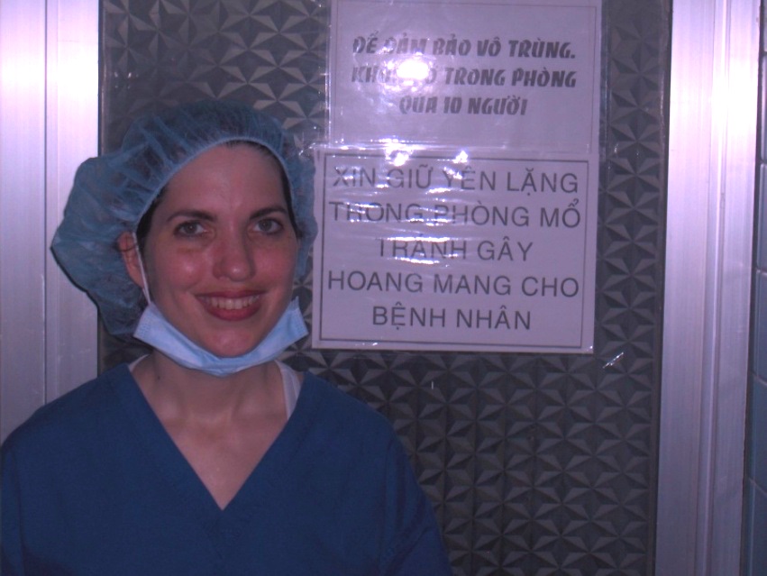 Woman in medical scrubs and cap smiling, standing next to a sign on a textured wall.
