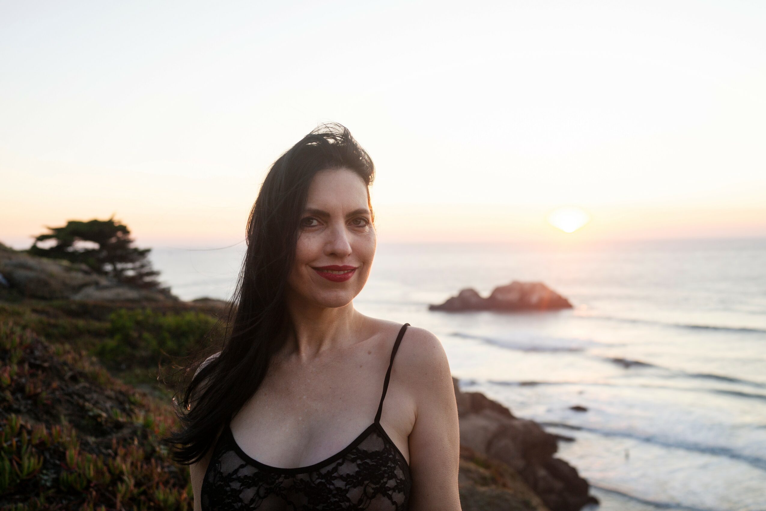 Woman with long dark hair standing outdoors near the coast at sunset, smiling, wearing a black lace top.