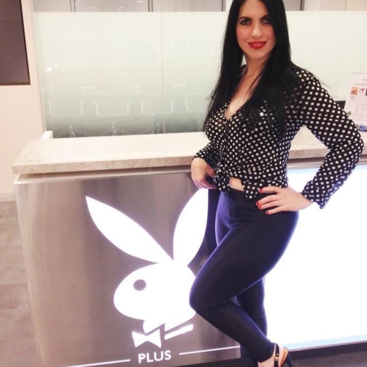 Woman with long dark hair, red lipstick, wearing a black polka dot top and dark pants, standing in front of a Playboy logo counter.