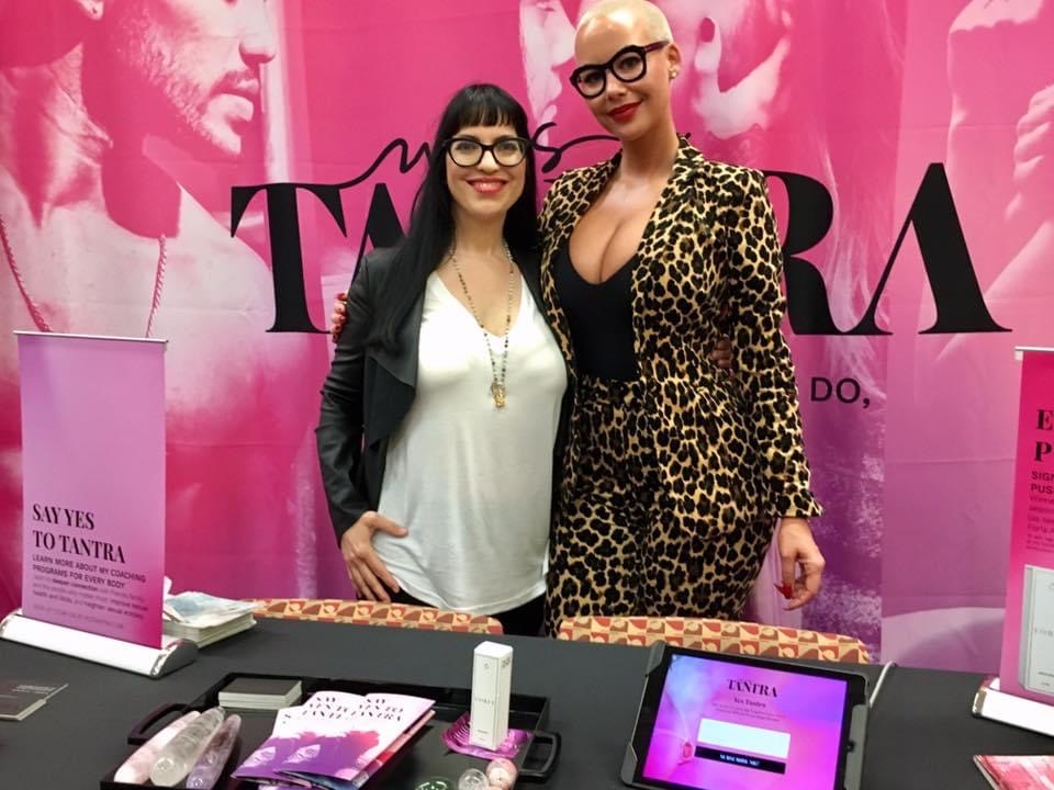 Two women standing in front of a pink backdrop with text, one wearing a leopard print outfit and glasses, the other in a black jacket and white top.