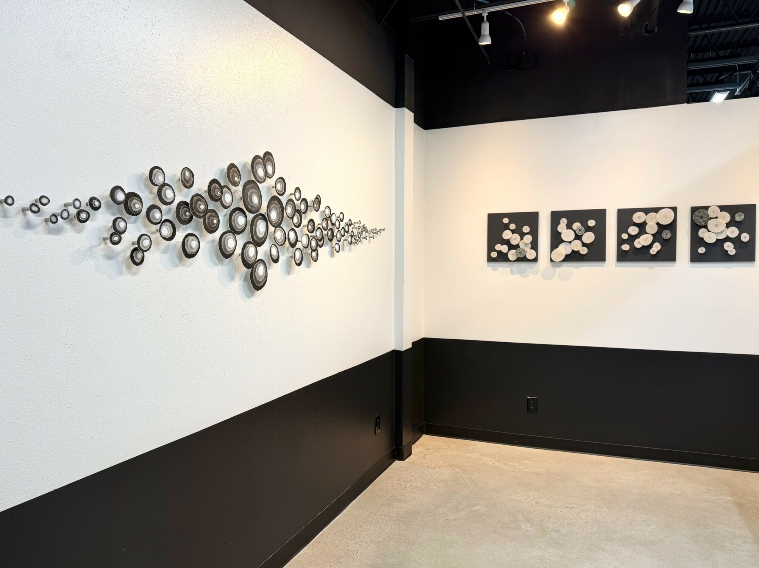 Art installation with circular objects on white wall, and four black-framed artworks with circular patterns, in gallery space.