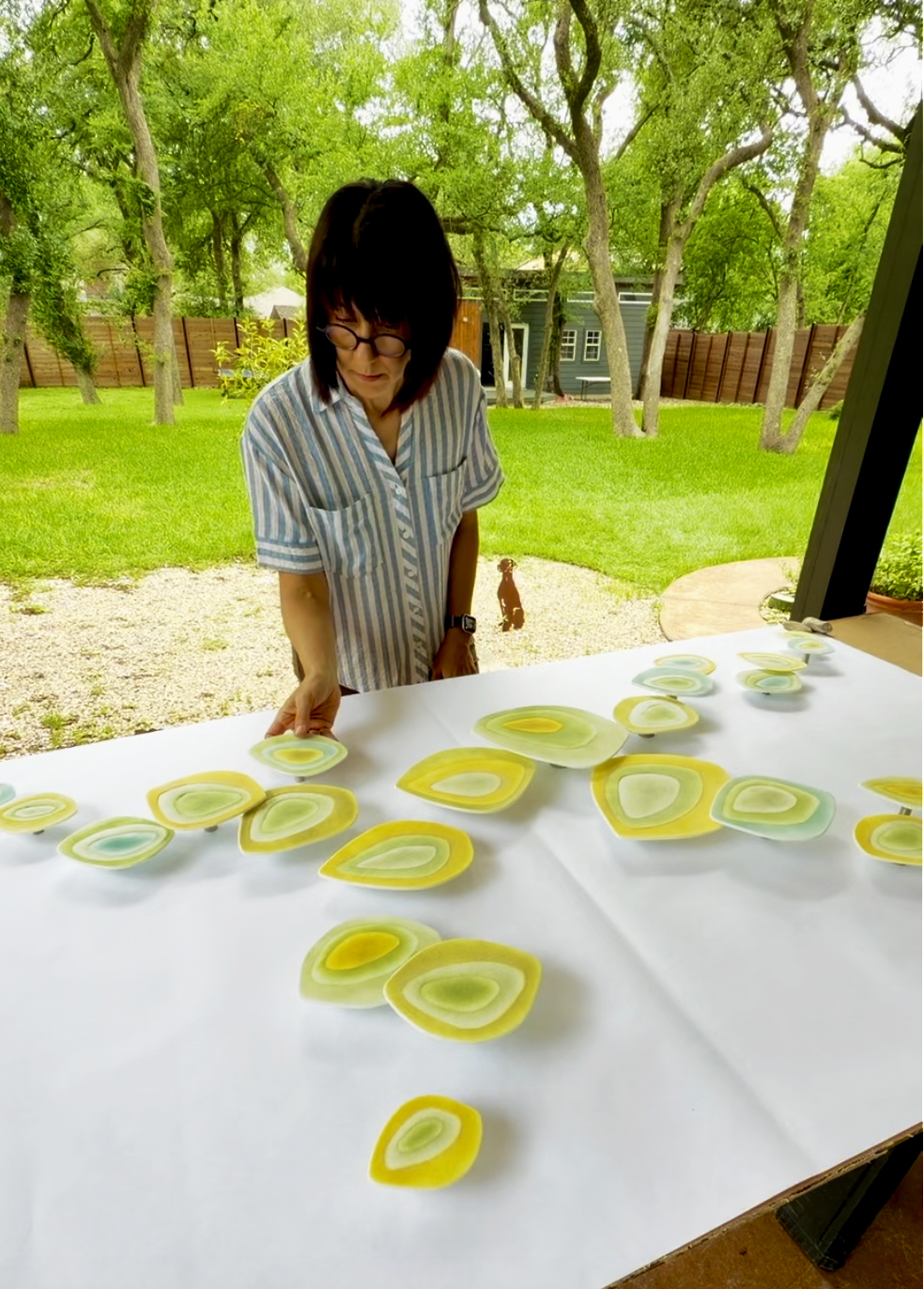 Person with dark hair and glasses reaching for yellow and green paper cutouts on a table outdoors, trees and grass in background.