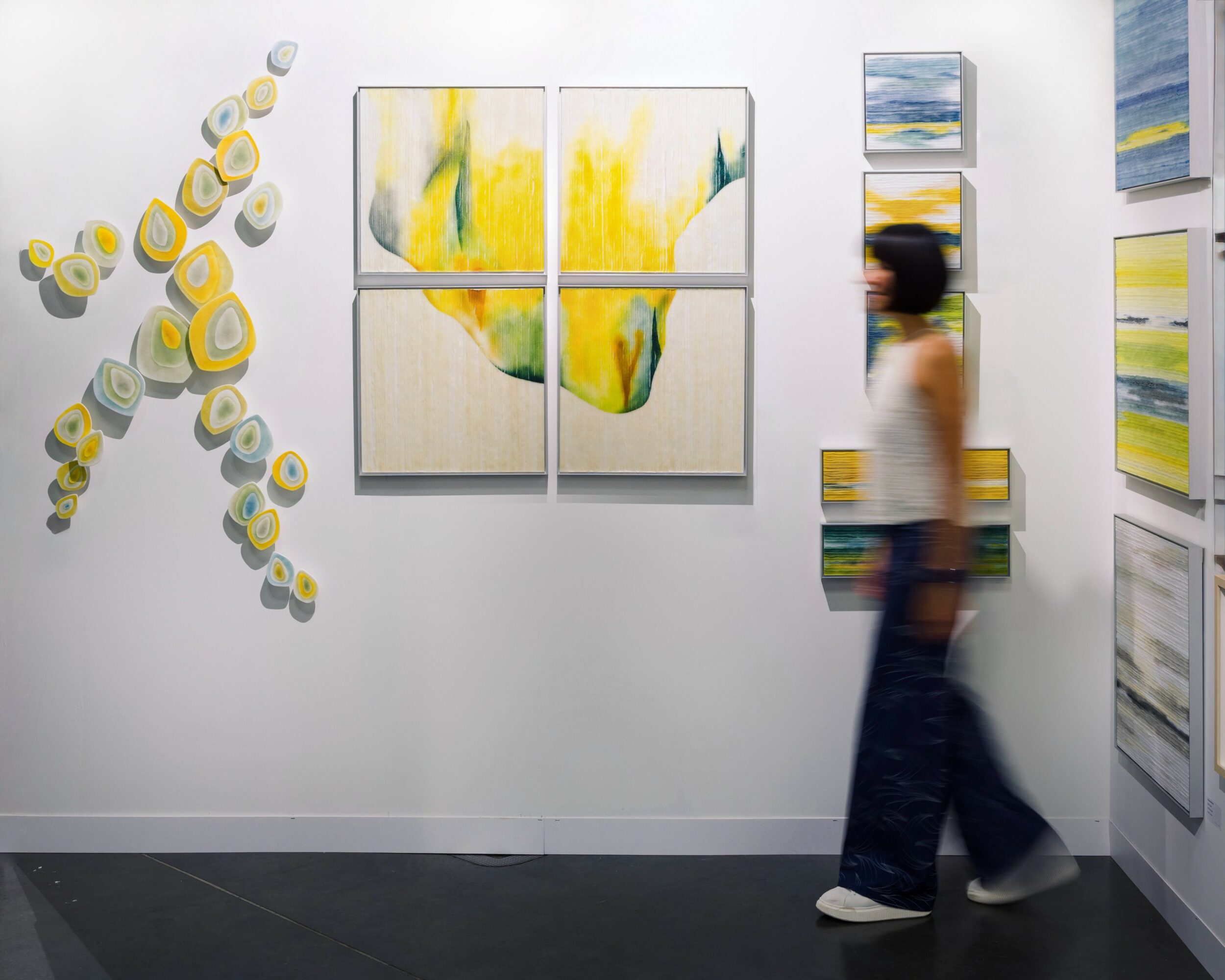Person walking past abstract paintings on gallery wall, with colorful circular and rectangular artwork, white background, black floor.