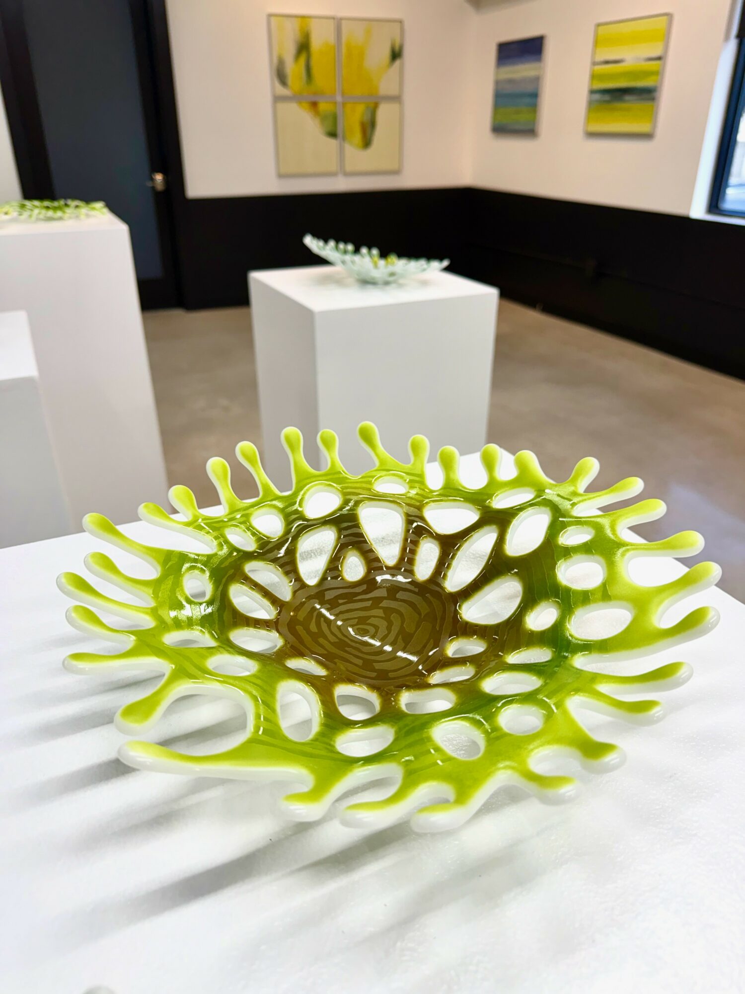 Decorative green and brown sculptural object with organic, openwork design on a white surface, art gallery background with paintings and display pedestals.