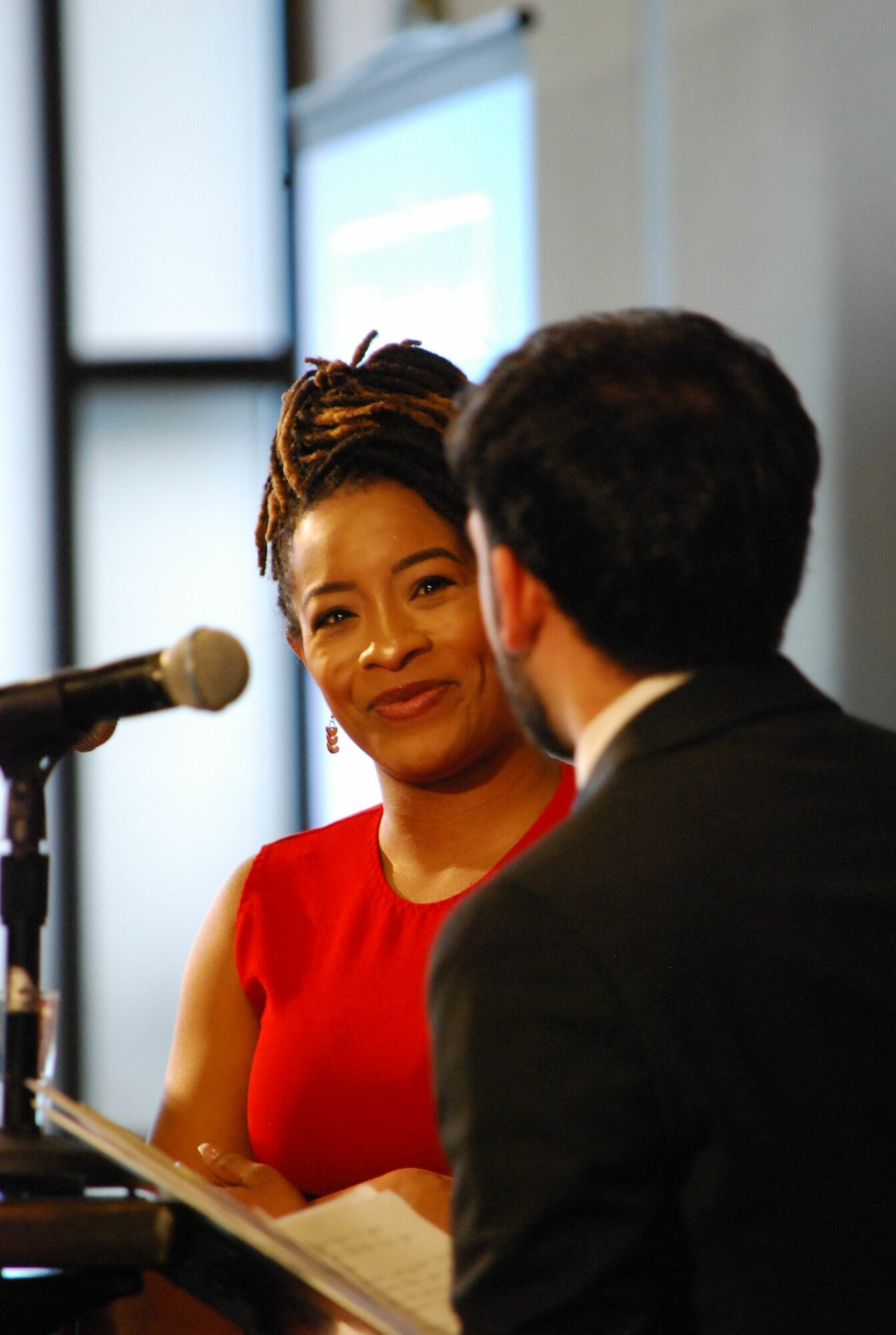 Woman in red dress speaking into a microphone, facing a man in a suit, in a professional setting.