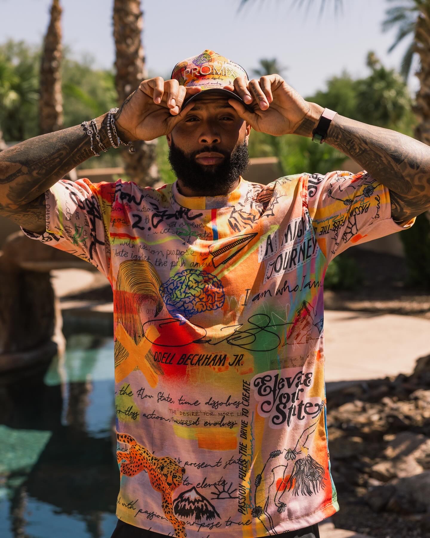 Man adjusting cap outdoors with trees and water in background, wearing a colorful graphic T-shirt, tattoos on arms, and a beard.