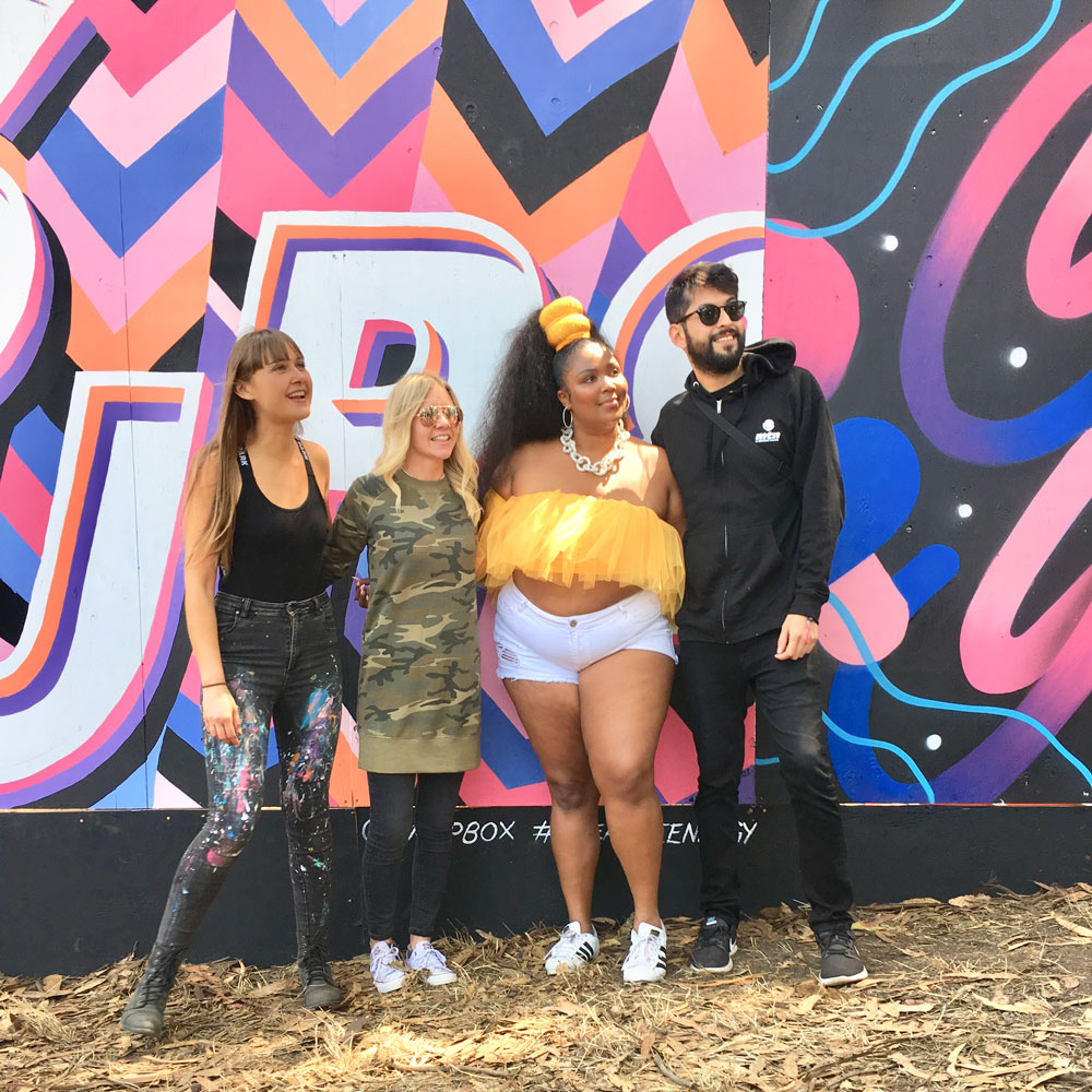 Four people standing in front of a colorful graffiti wall, smiling and posing outdoors on dirt ground.