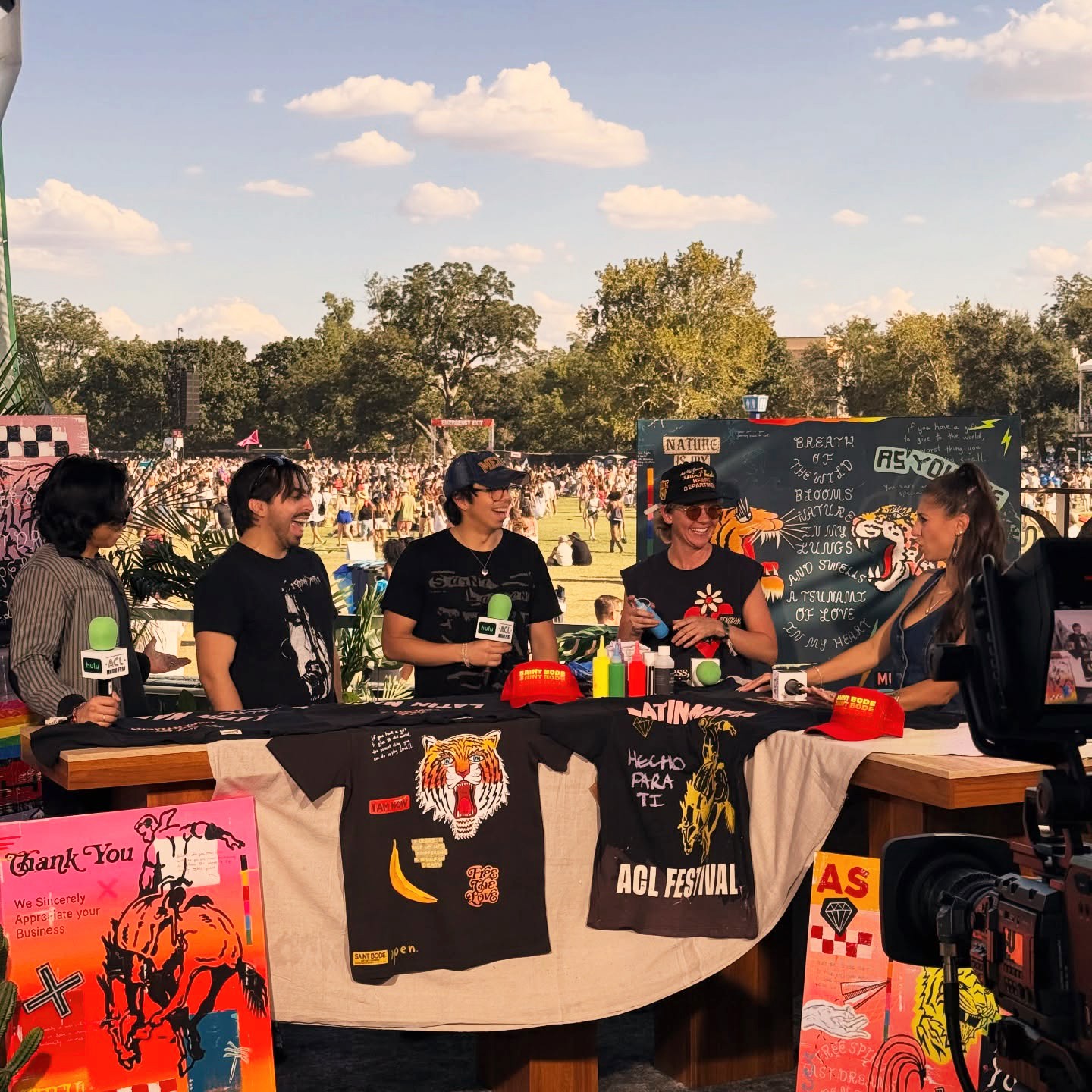 Group of five people at a booth outdoors, with trees and a crowd in the background, exchanging items or information.