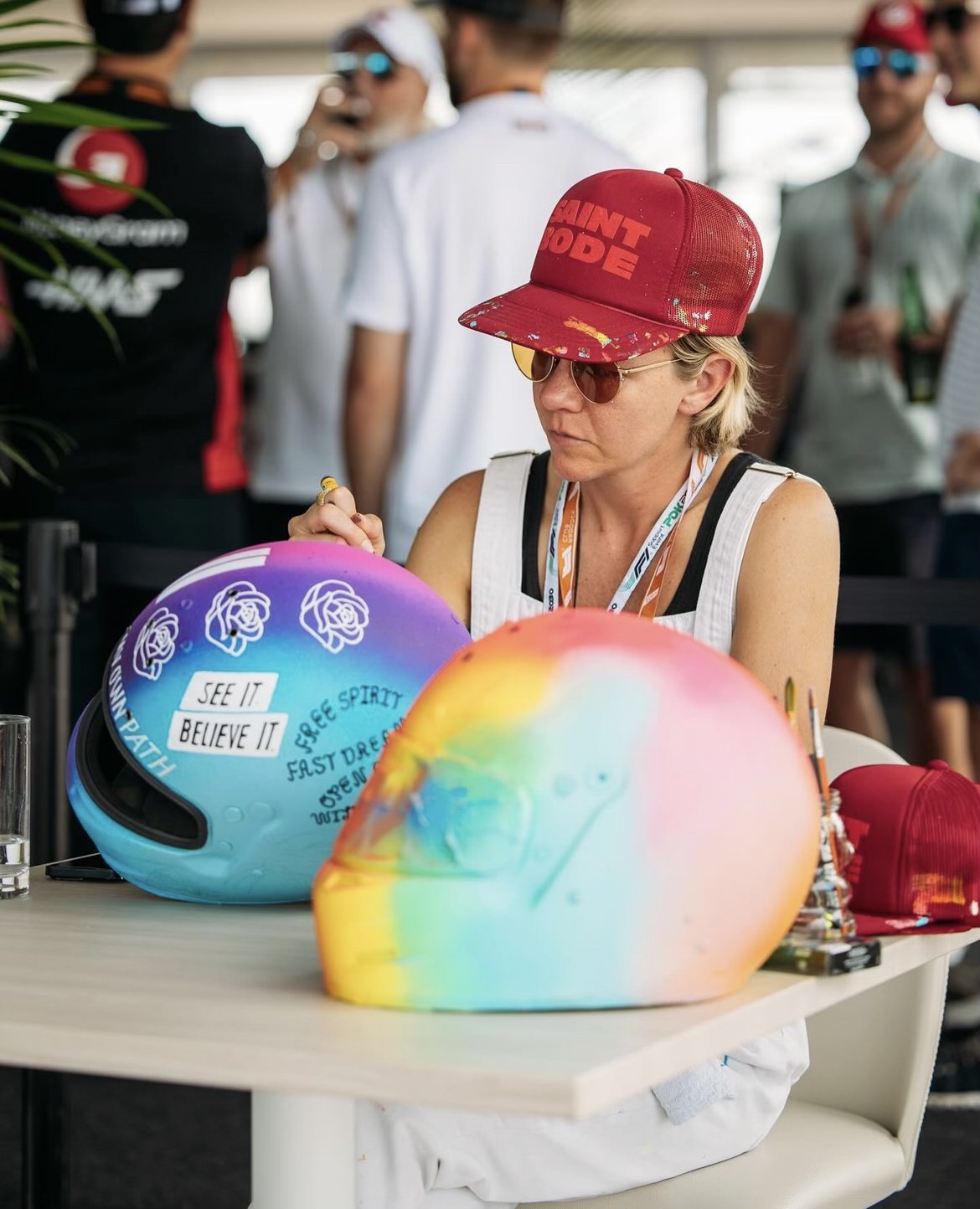 Woman wearing a red cap and sunglasses at a table with colorful helmets, in a crowded outdoor setting.