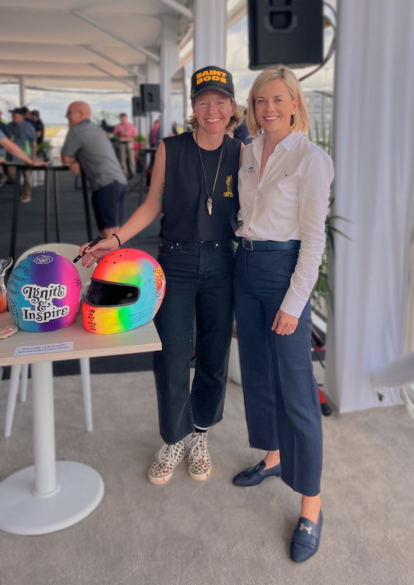 Two women standing indoors, smiling, with a table and colorful helmet in front of them.