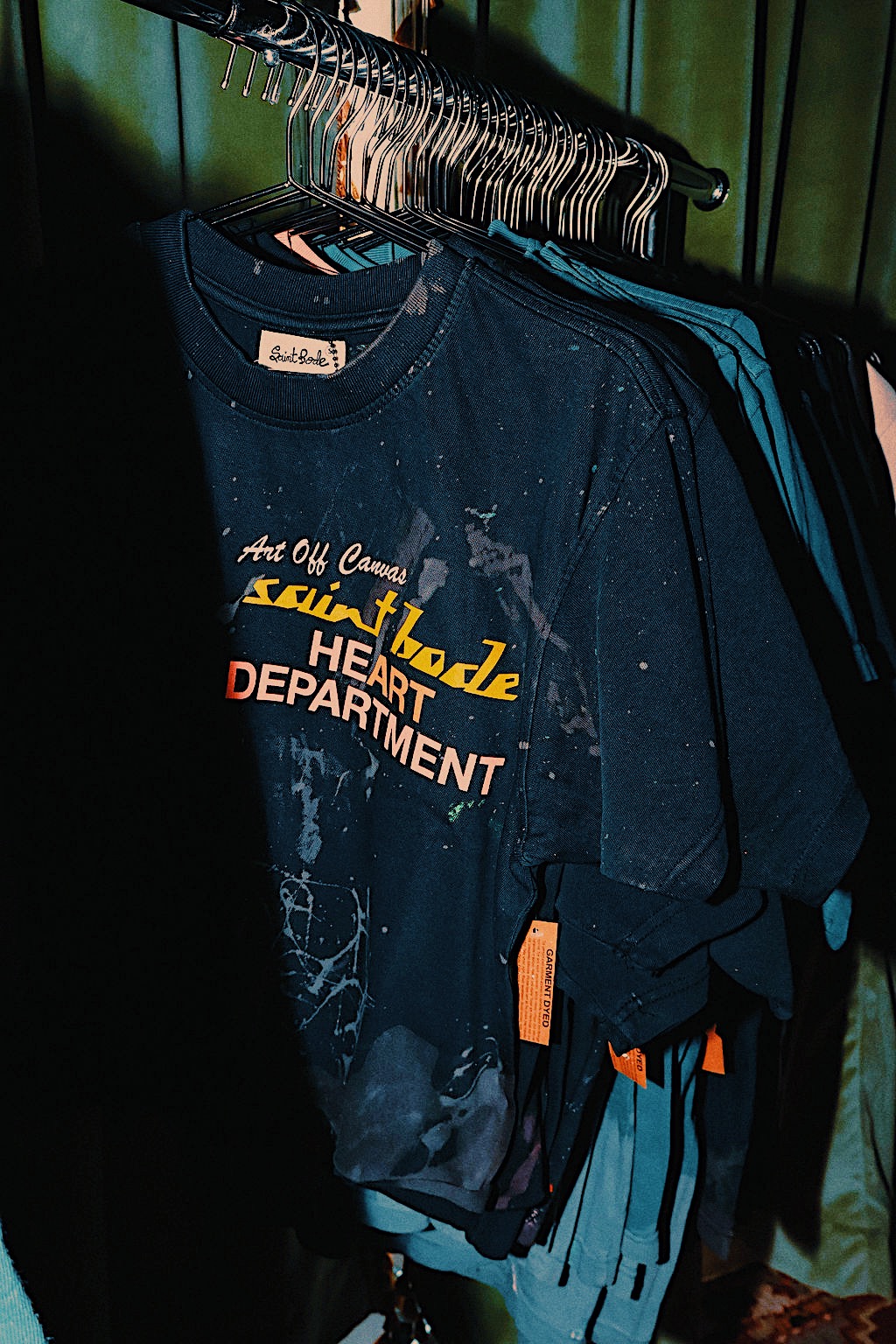 Dark blue T-shirt with yellow and white text hanging on a rack among other clothes, against a green curtain background.