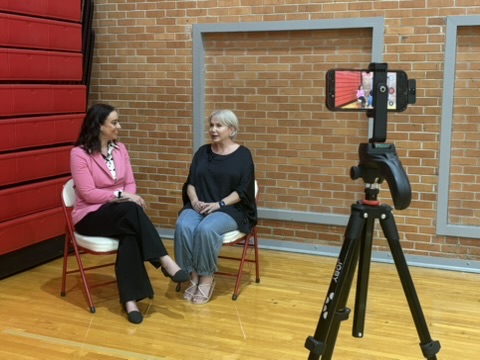 Two women sit on chairs in a room with a brick wall, recording a video with a camera on a tripod.