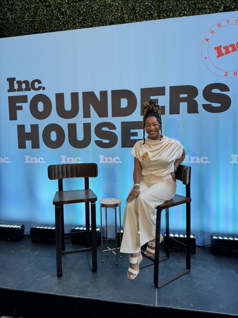 Woman sitting on a high chair on stage with a blue backdrop and two empty chairs, smiling, wearing a white dress.