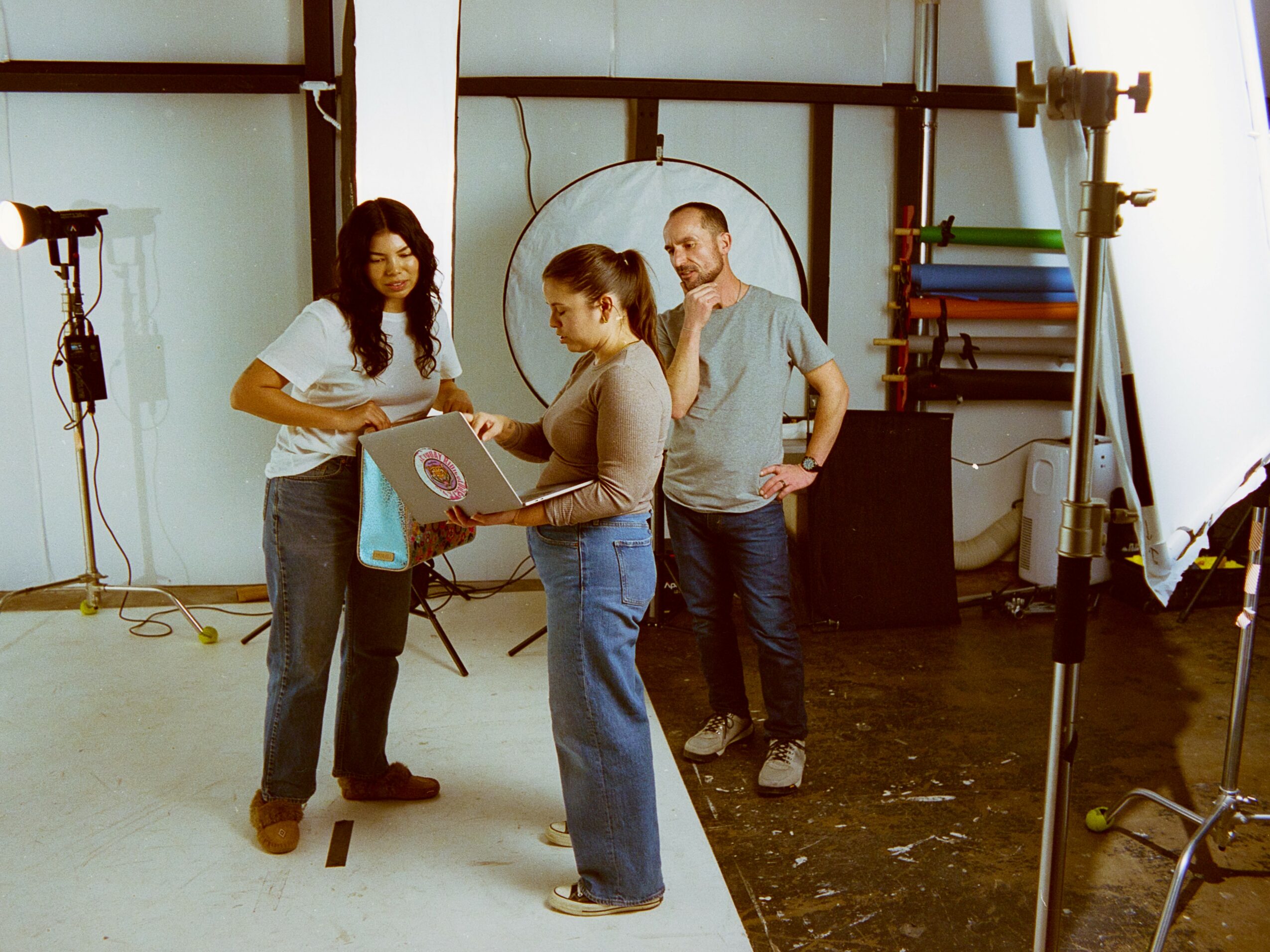Three people in a photography studio discussing a laptop, with photography equipment and a backdrop in the background.