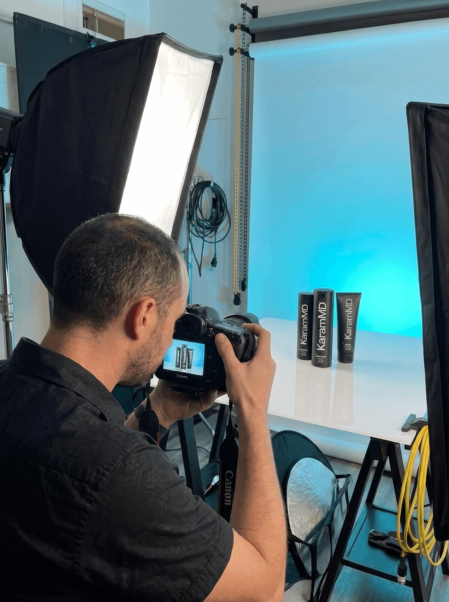 Photographer taking picture in studio with lighting and backdrop, camera view visible, person with short hair wearing black shirt.