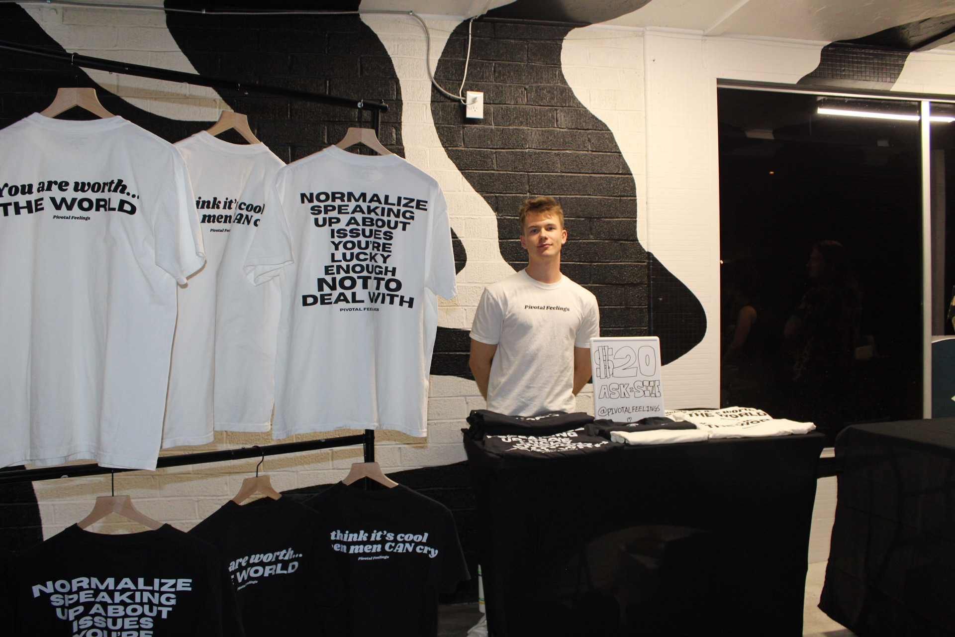 Young person standing behind a table with T-shirts, in a room with black and white wall art, and a TV screen.