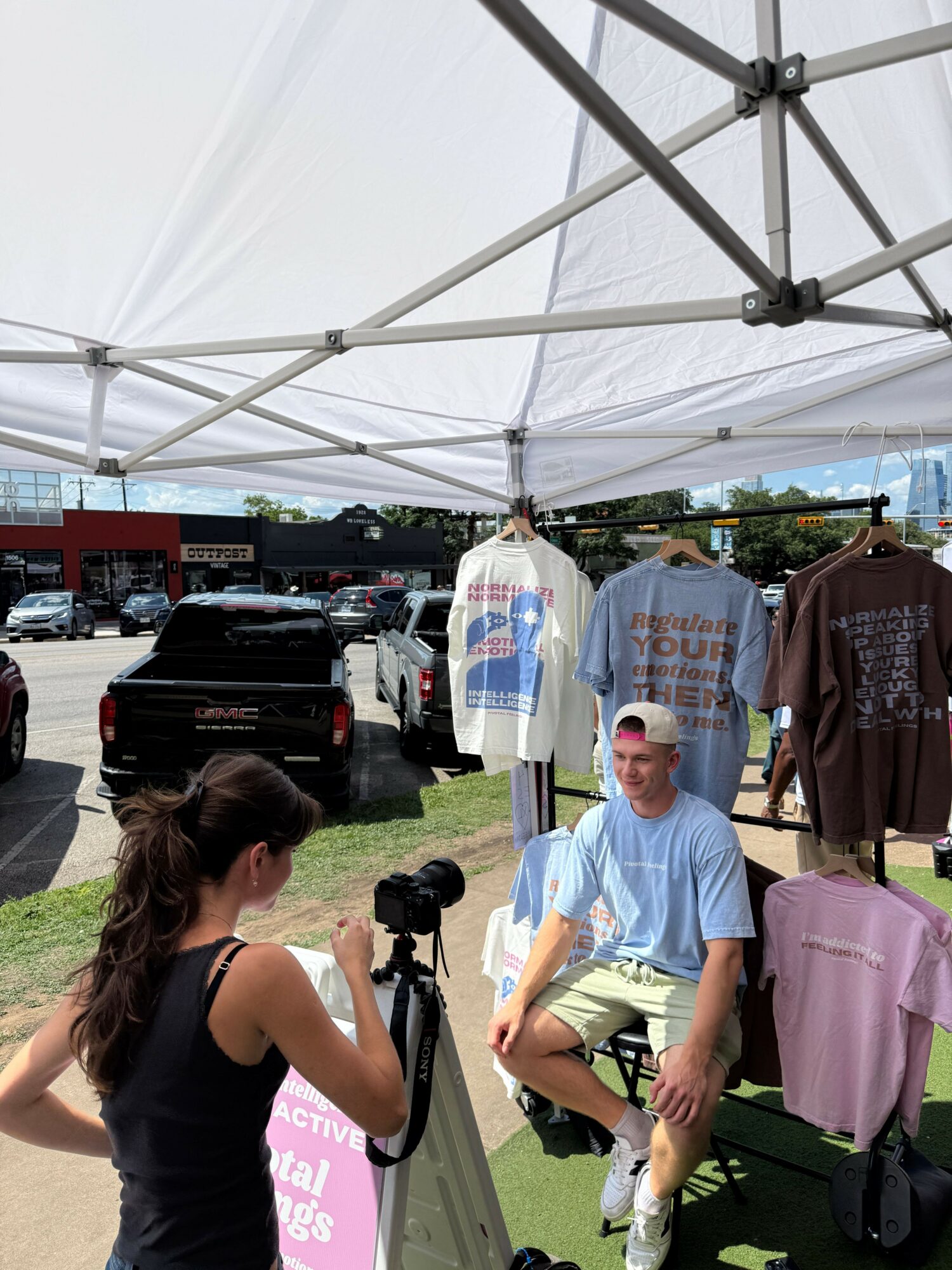 Man in light blue shirt and beige shorts standing under a white canopy tent, with clothing and bags displayed behind him, outdoors.