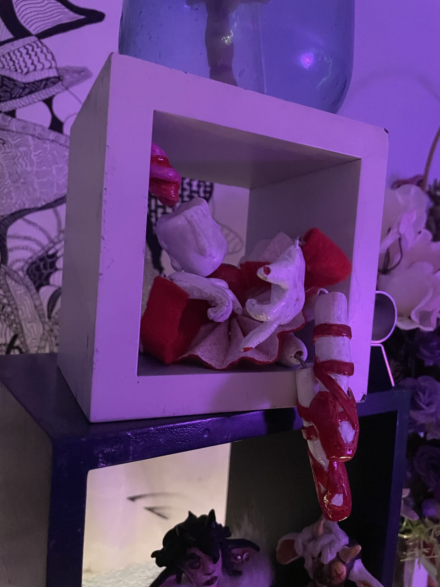 Decorative doll inside a white square frame, with a purple background and flowers nearby.