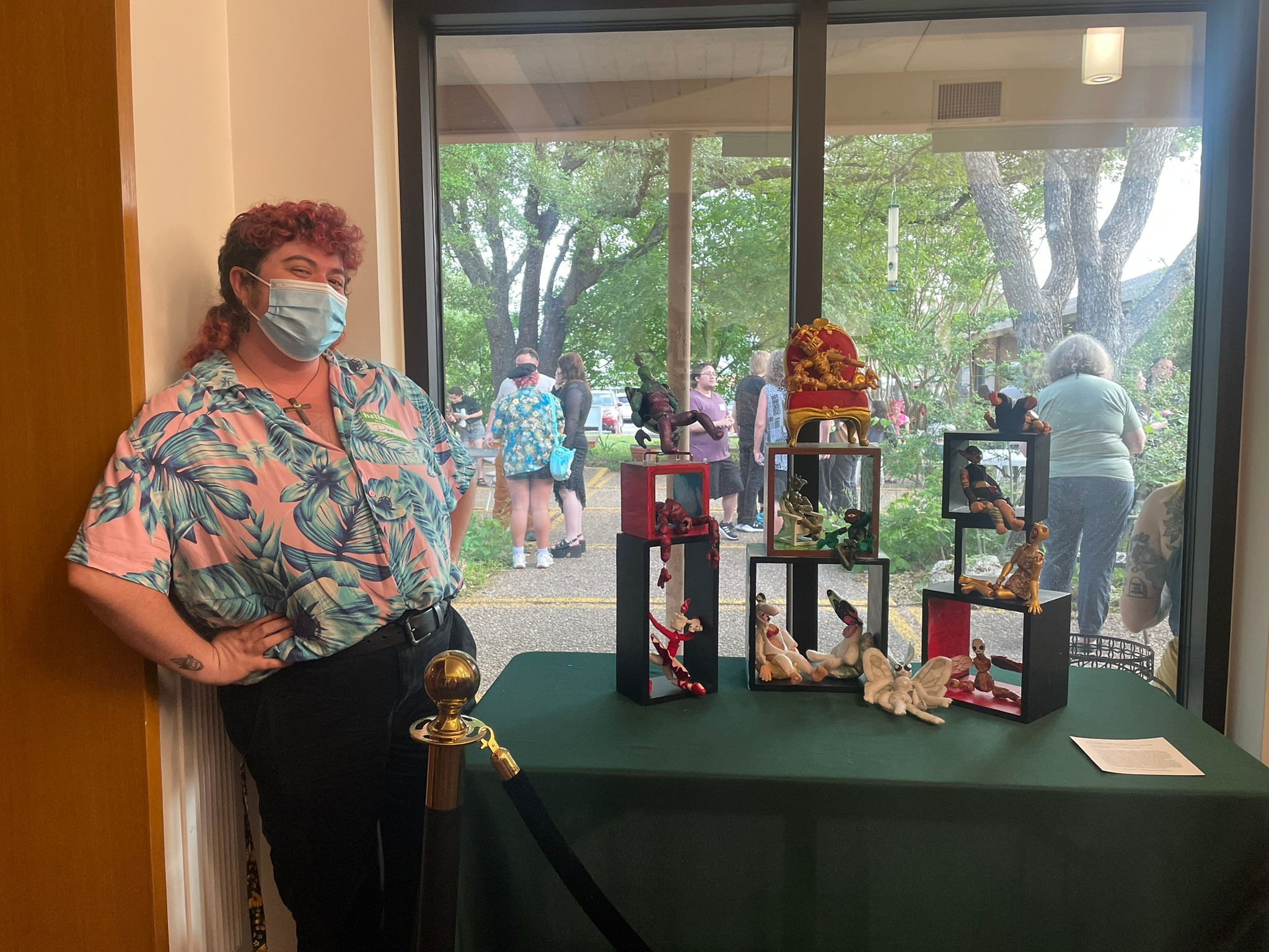 Person wearing a face mask and colorful shirt standing next to a display table with framed photos and decorations, outside window view.