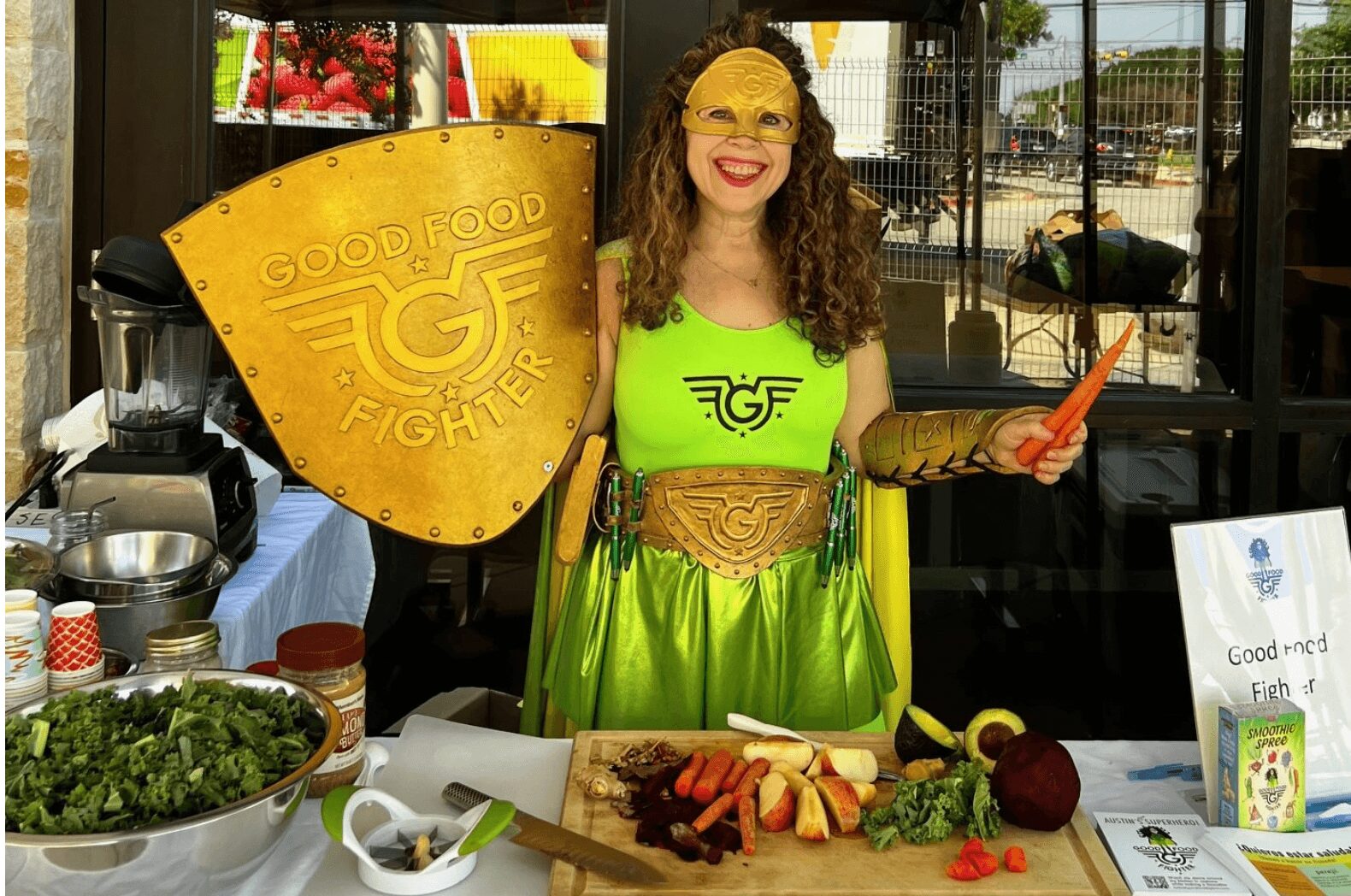 Person dressed as a food fighter holding a shield and a spatula, smiling, standing behind a table with food, in a bright setting.