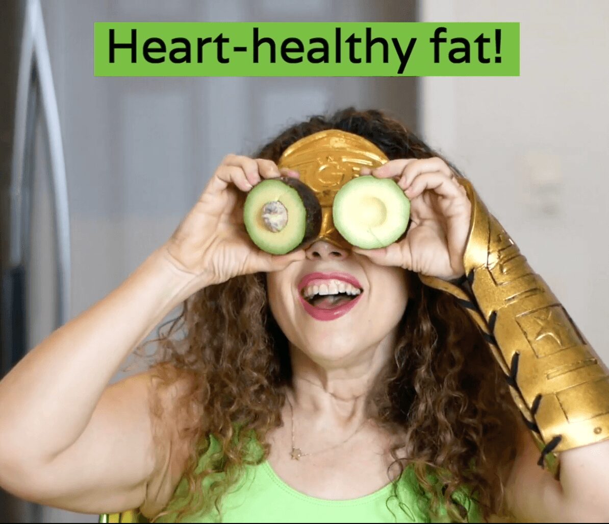 Woman with curly hair holds kiwi and cucumber slices over eyes, smiling, with a green text overlay reading 'Heart-healthy fat!'.