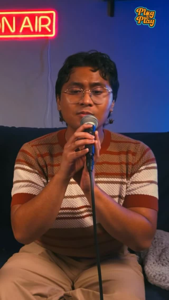 Person with glasses holding a microphone, sitting in front of a blue background with a neon 'ON AIR' sign, and a small logo.