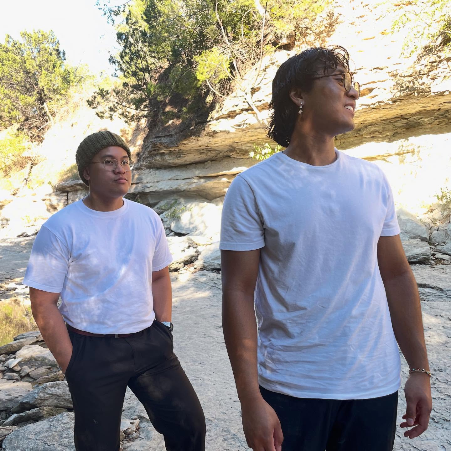 Two people standing outdoors near rocks and trees, both wearing white t-shirts and dark pants, one with hands in pockets.