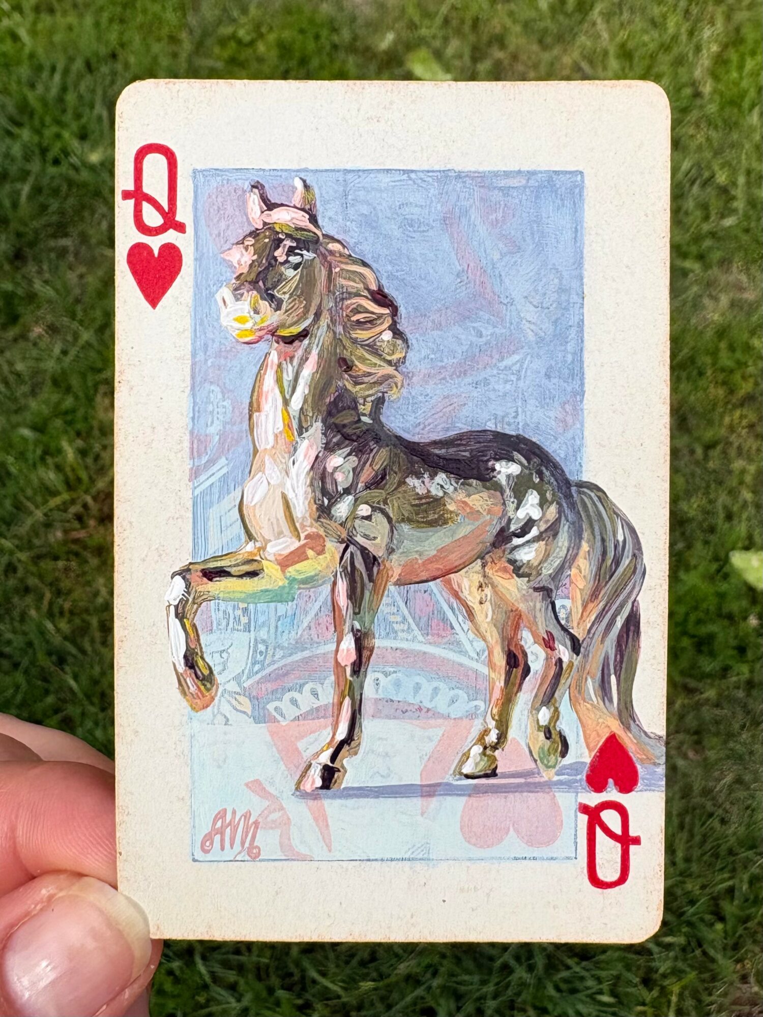Playing card with a horse illustration, red heart symbols, and a white border, held outdoors with grass background.
