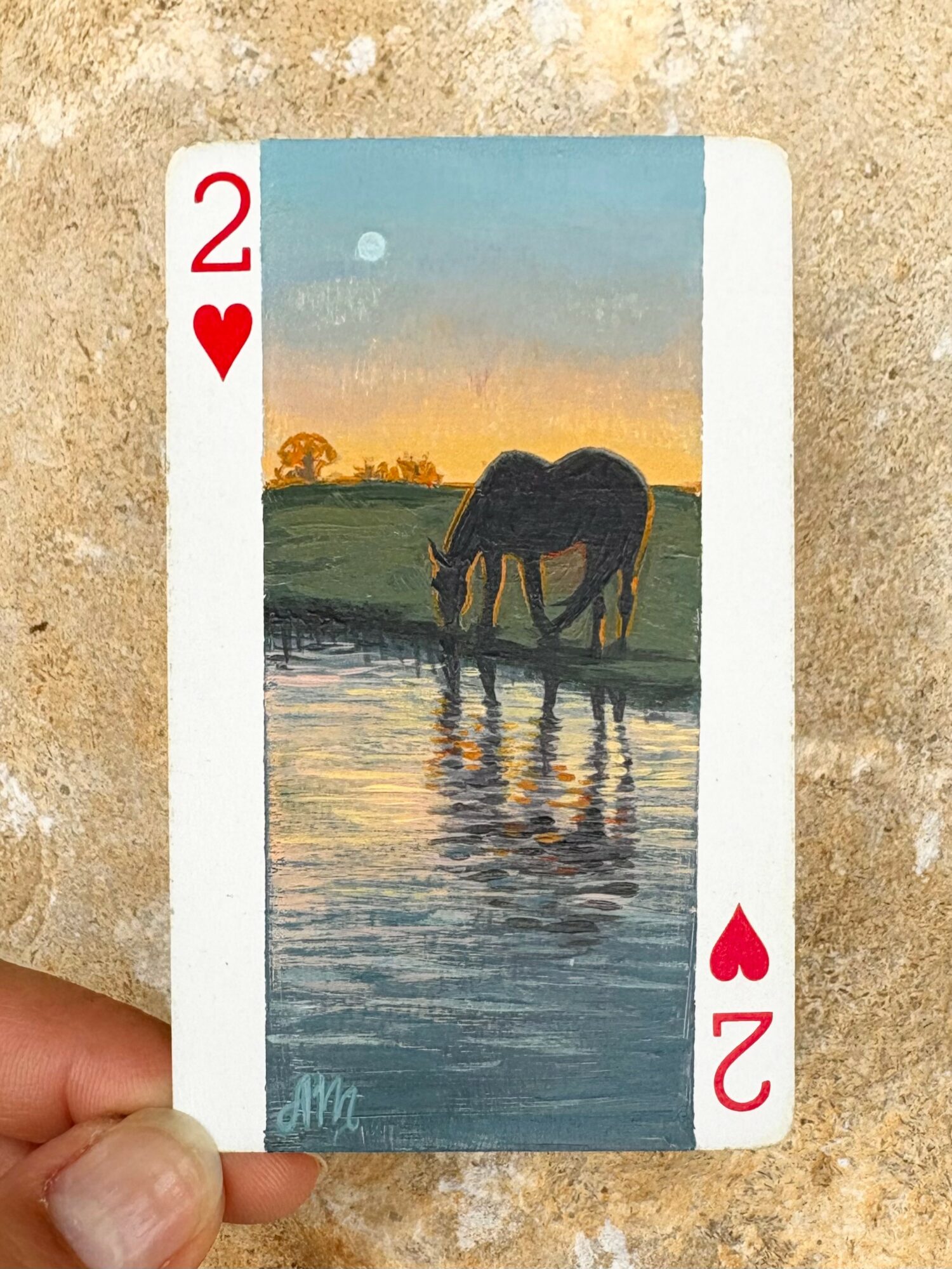 Playing card with a horse drinking water at sunset, landscape background, red hearts, number 2, and a white border.
