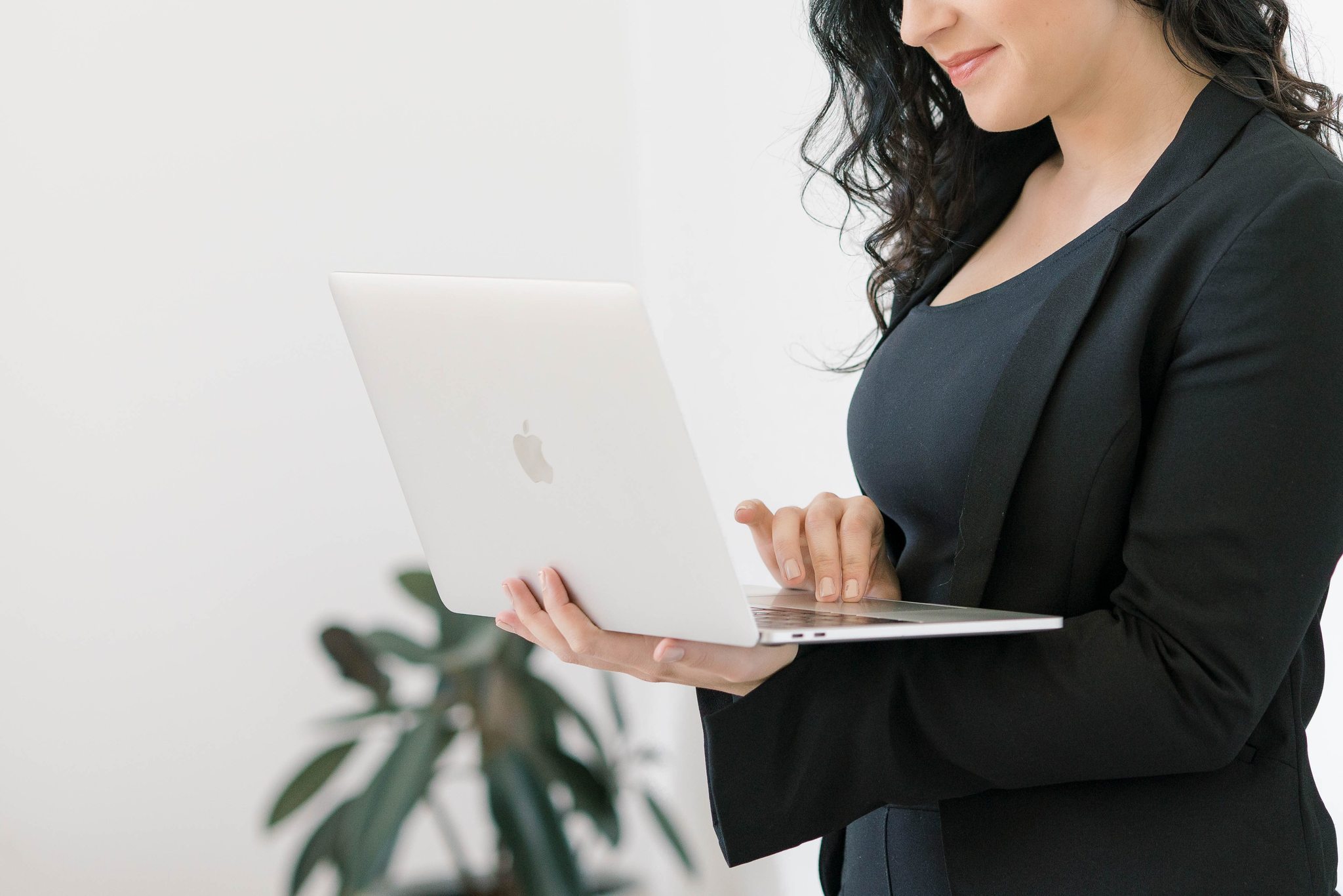 Woman in black blazer using a laptop, with a plant in the foreground, against a white background.