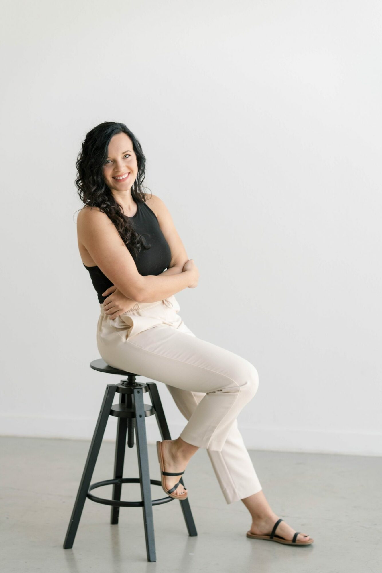 Woman with curly hair sitting on a stool, smiling, wearing a black top and white pants, against a plain background.