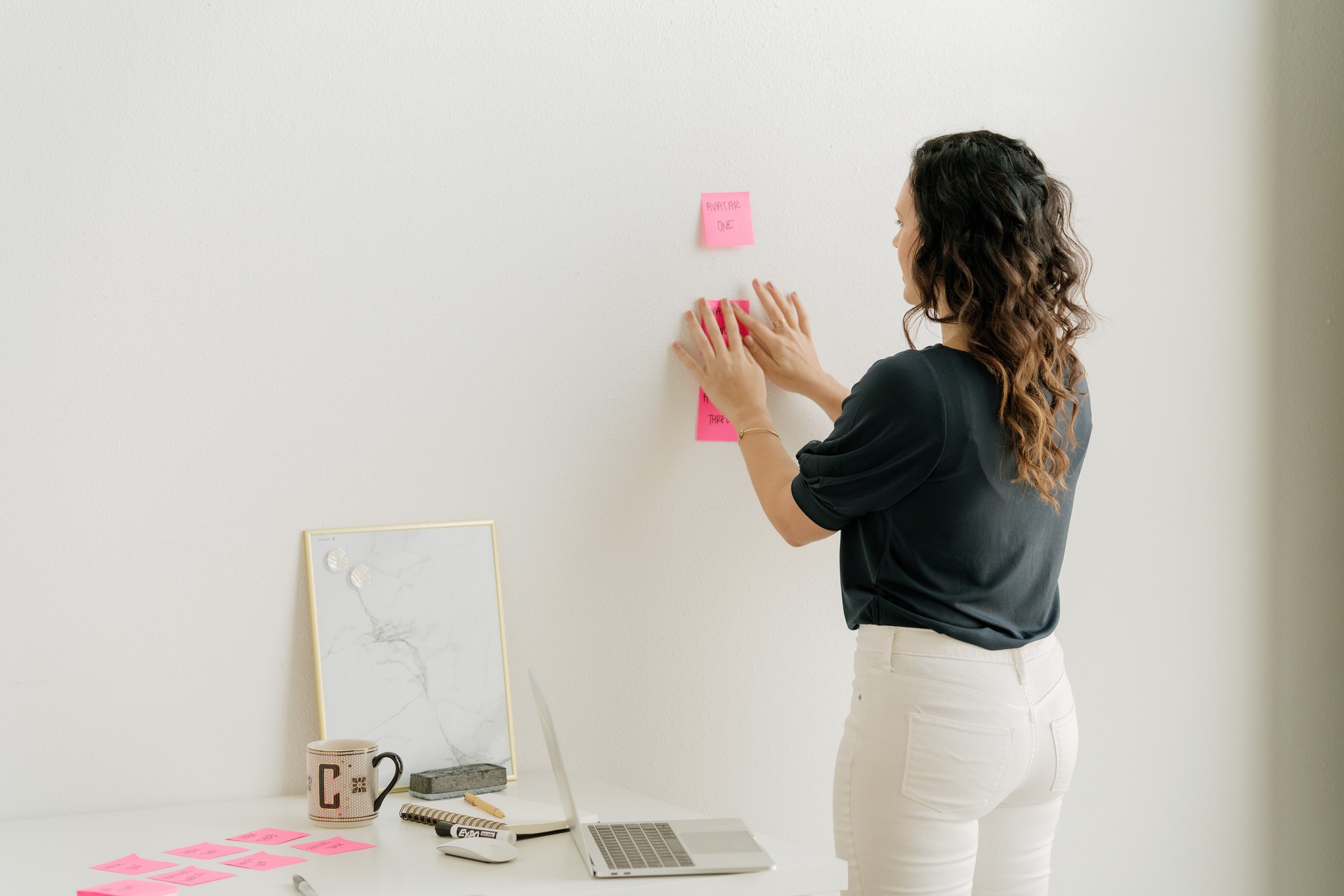 Woman with curly hair in black top and white pants sticks pink notes on a white wall, with a desk and laptop nearby.
