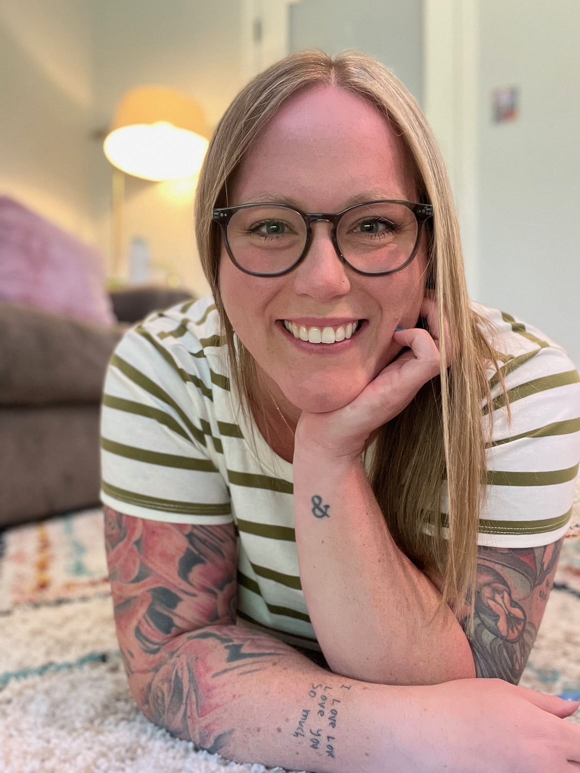 Woman with glasses smiling, resting her chin on her hand, wearing a striped shirt, tattoos on arms, in a cozy room.